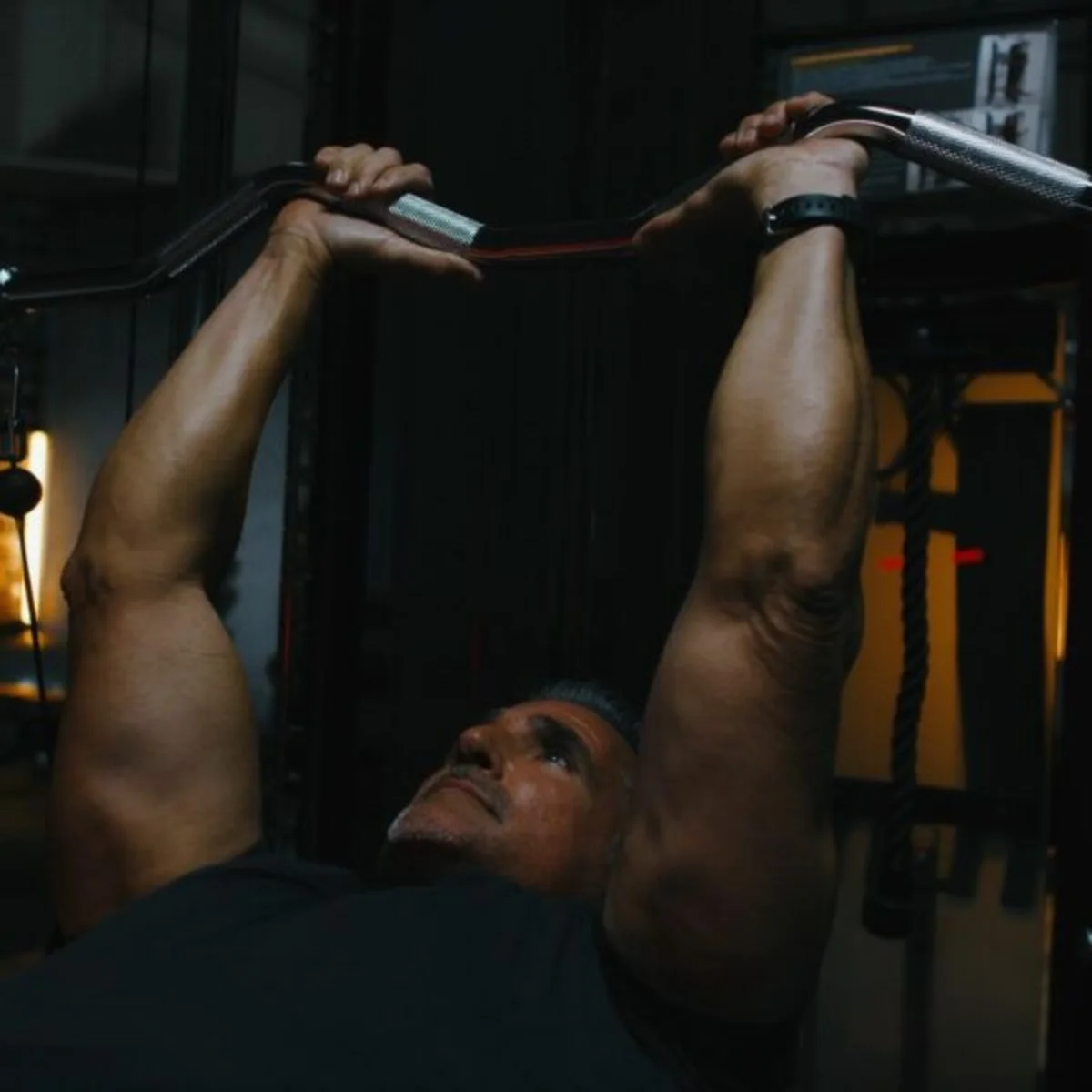 Man in gym, lifting barbell over chest, looking upwards, focused.