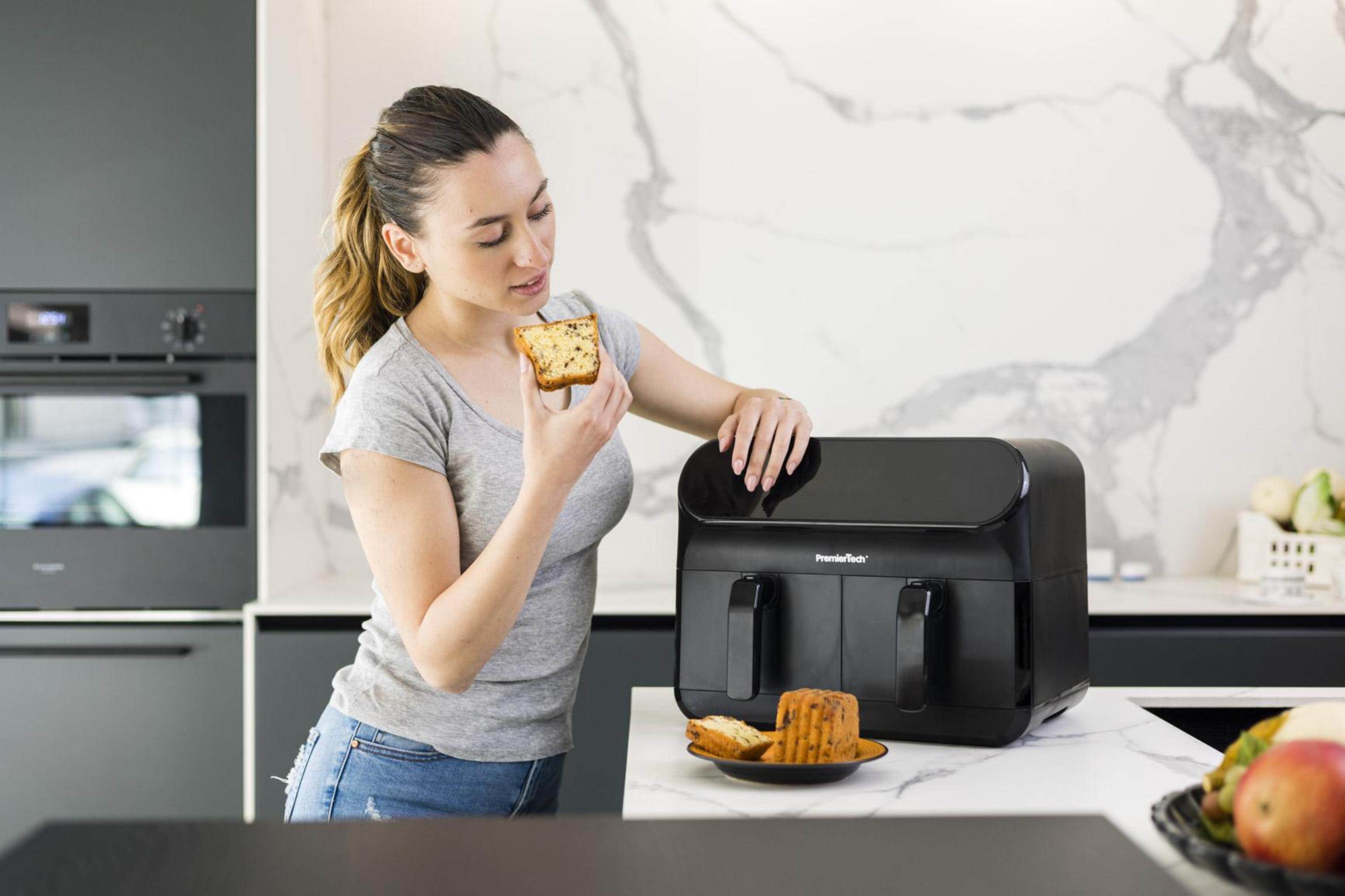 Donna che tiene in mano una fetta di torta vicino a una friggitrice ad aria nera, con cibo sul bancone e sullo sfondo.