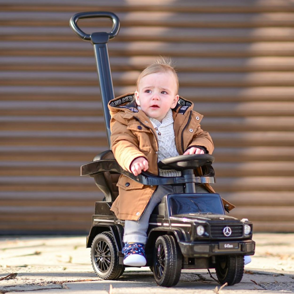 Ein Kleinkind in einem Spielzeugauto, trägt eine braune Jacke und schaut hoch.