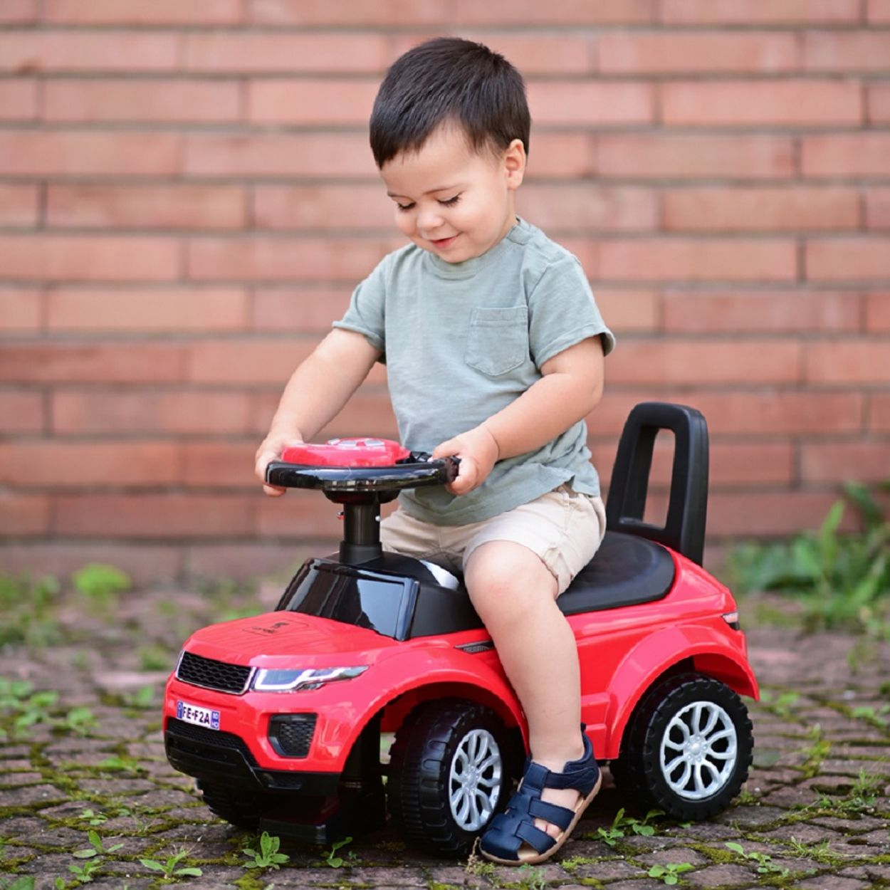 Un niño en un coche de juguete rojo. Fondo de pared de ladrillo. El niño sonríe.