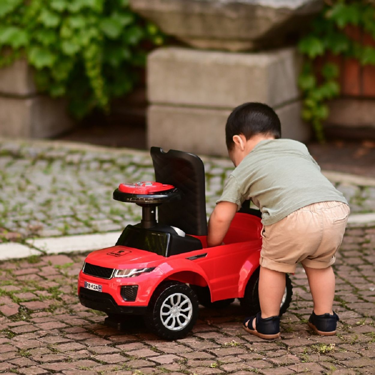 Un niño pequeño se inclina hacia un coche de juguete rojo, en un área exterior pavimentada con ladrillos.