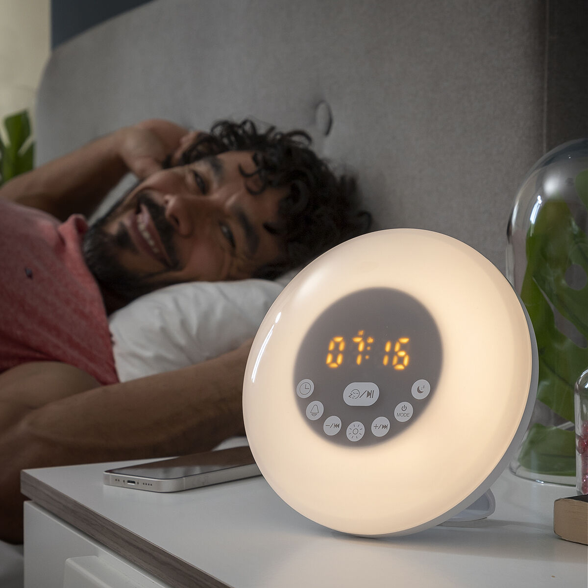 Un hombre sonriendo en la cama con un despertador brillante en la mesita de noche.