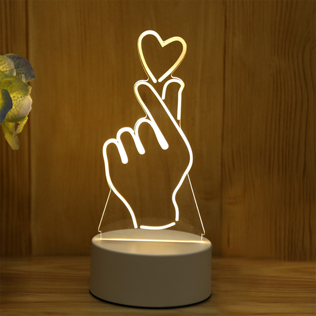 Eine handförmige LED-Lampe mit Herzgeste, auf weißem Sockel, mit Holz-Hintergrund.