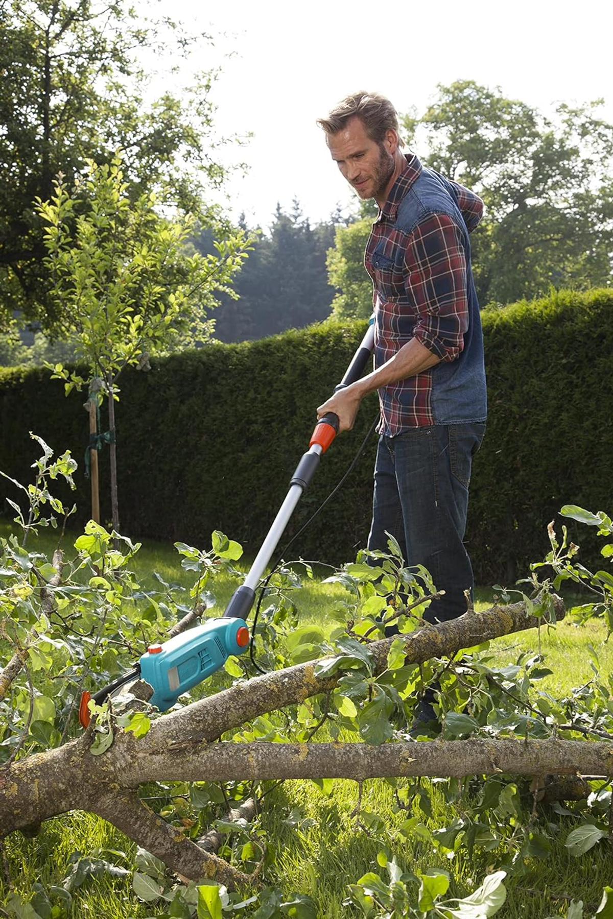 Un uomo che taglia un ramo con una sega elettrica. È in un giardino con erba verde.