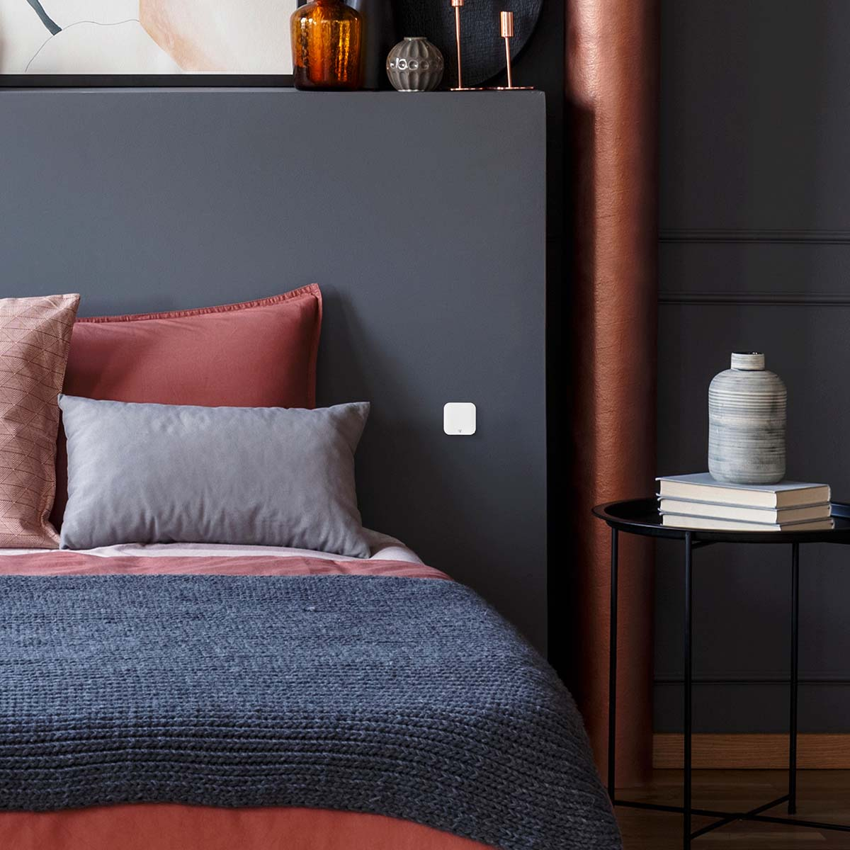 Slaapkamer met bed. Rode en grijze kussens, blauw deken. Muur heeft lichtschakelaar. Nachtkastje met objecten.