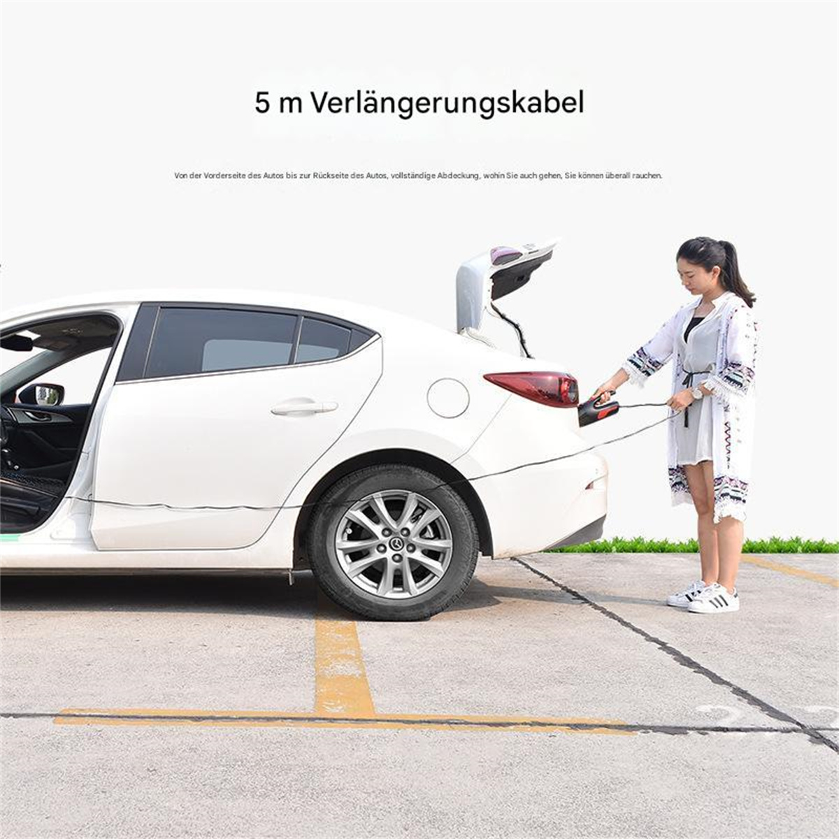 Eine Frau verbindet ein schwarzes Kabel mit dem offenen Kofferraum eines weißen Autos auf grauem Untergrund.