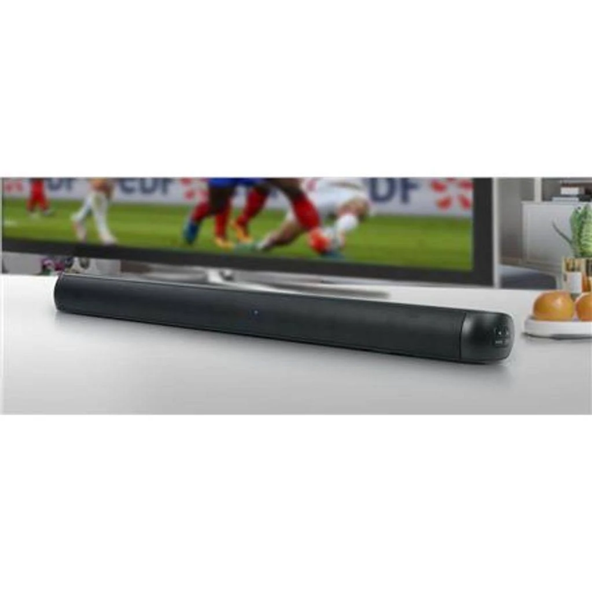 Soundbar nera su una superficie bianca. Schermo TV sfocato con partita di calcio sullo sfondo.