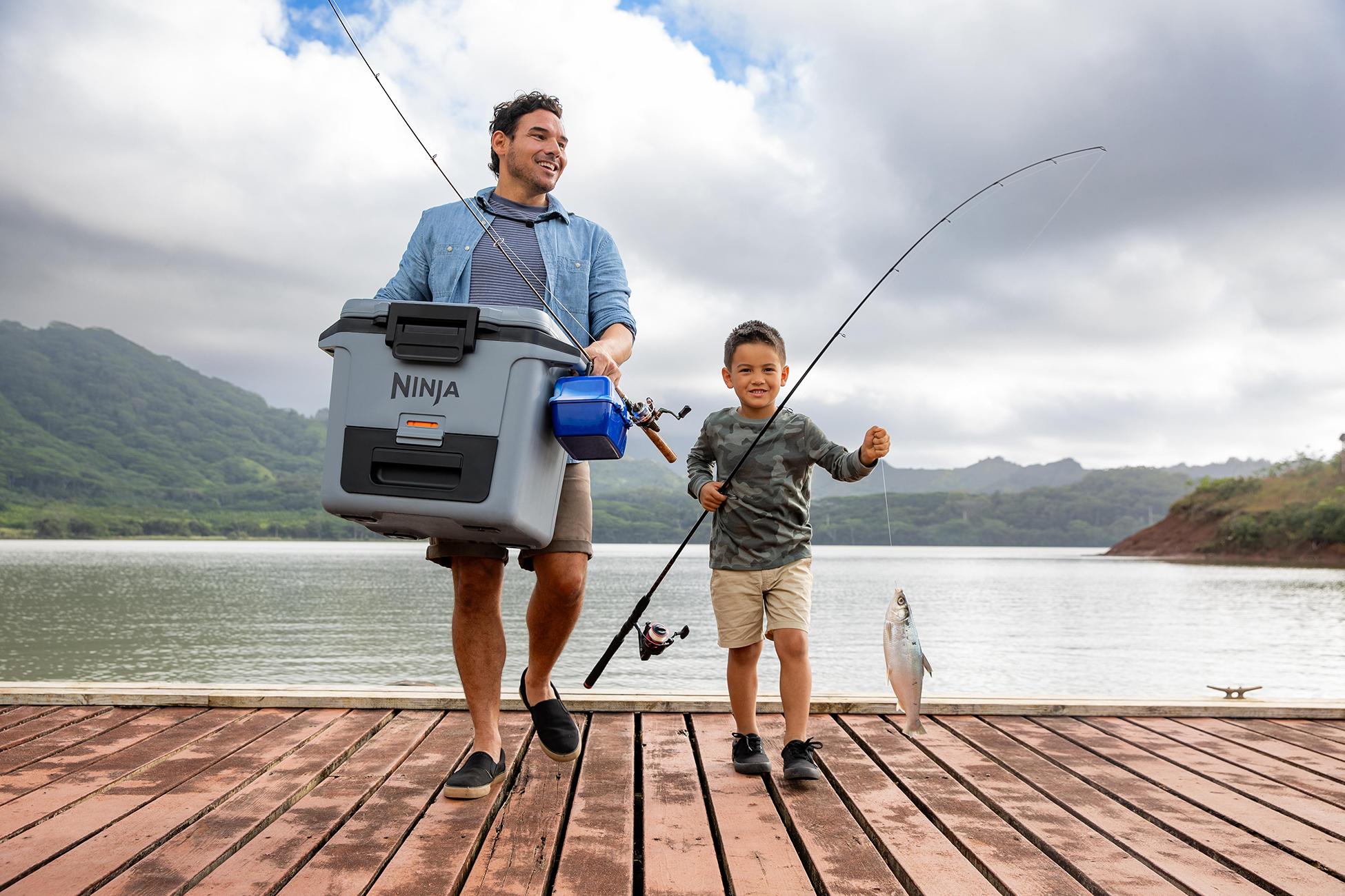 Ojciec i syn idący po pomoście z wędkami i lodówką. Jezioro i niebo w tle.