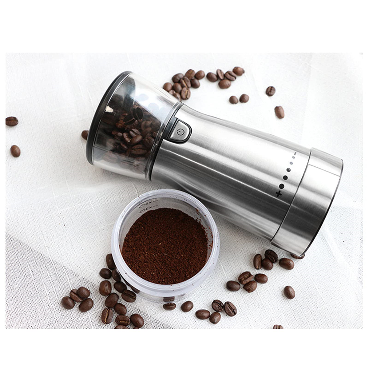Silberne elektrische Kaffeemühle mit Bohnen und gemahlenem Kaffee auf weißem Tuch.