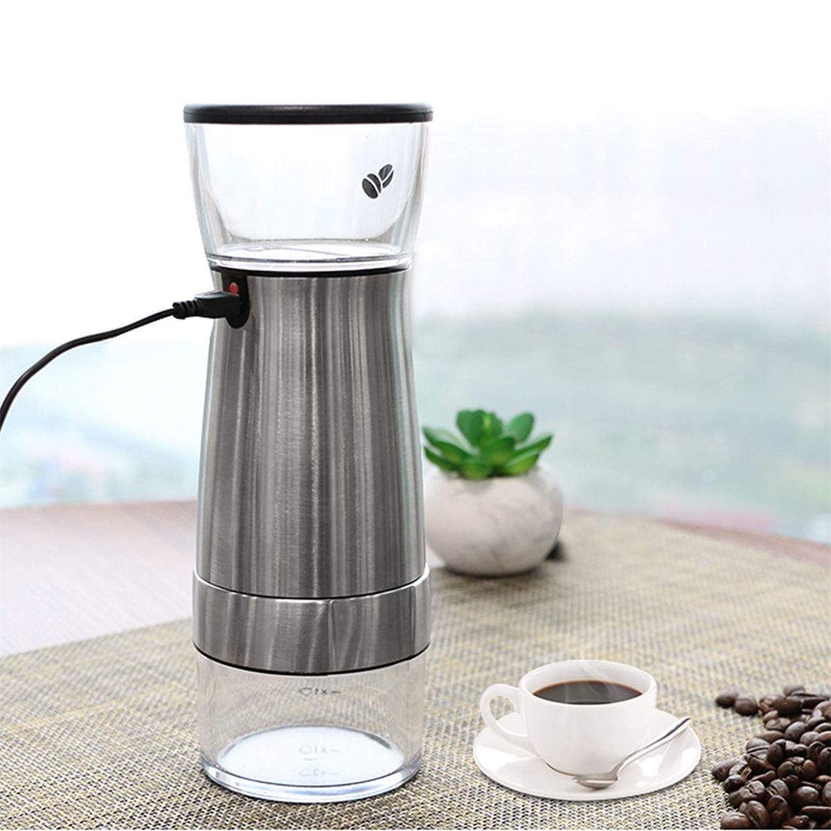 Silberne elektrische Kaffeemühle. Kaffeetasse auf Untertasse mit Kaffeebohnen auf dem Tisch.