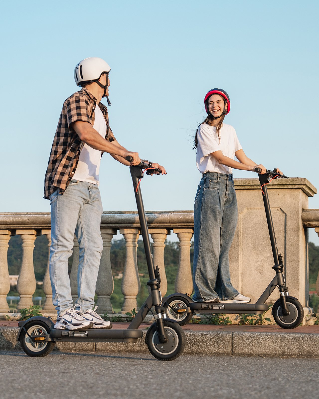 Zwei Personen fahren Roller. Sie lächeln und tragen Helme. Der Hintergrund ist ein blauer Himmel.
