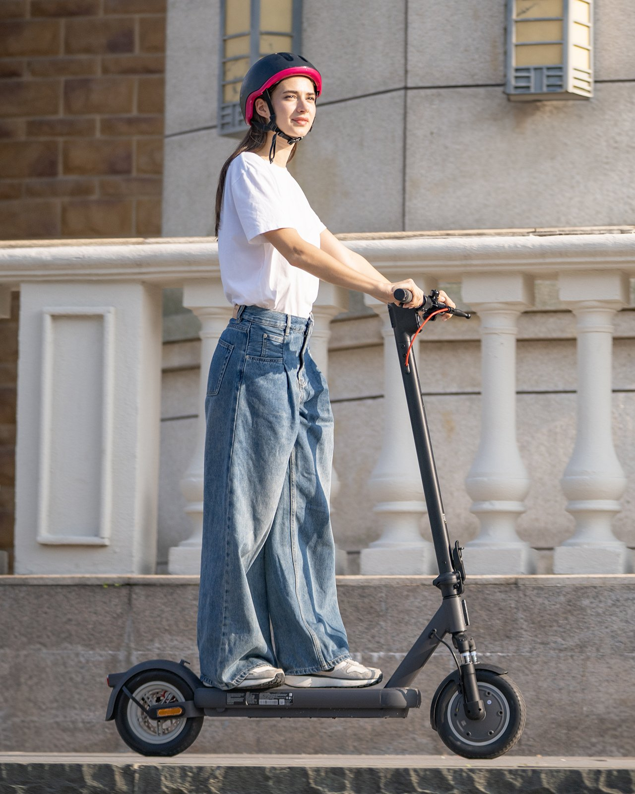 Frau auf einem Roller mit Helm, steht auf einem Bürgersteig mit Gebäude und Geländer.
