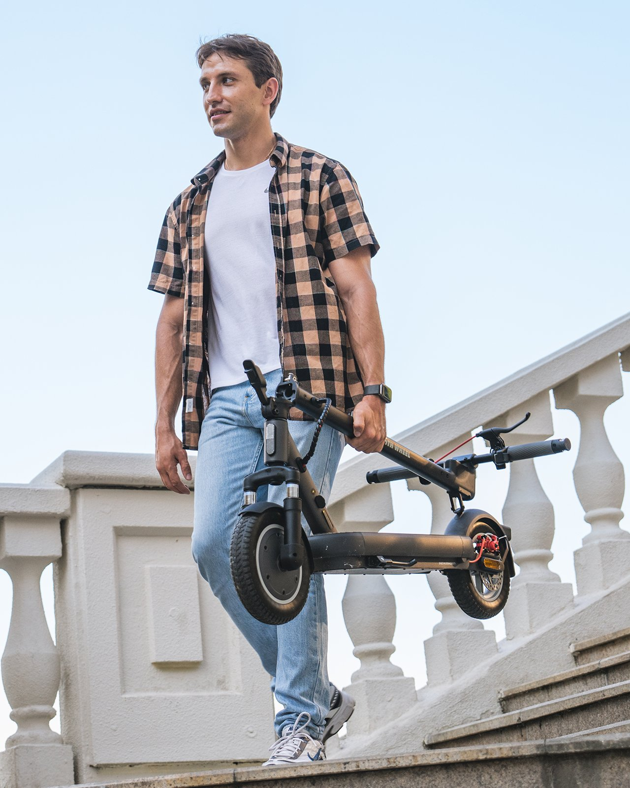 Mann trägt zusammengeklappten schwarzen Elektroroller. Er geht Treppen hinunter, trägt ein Hemd und Jeans.