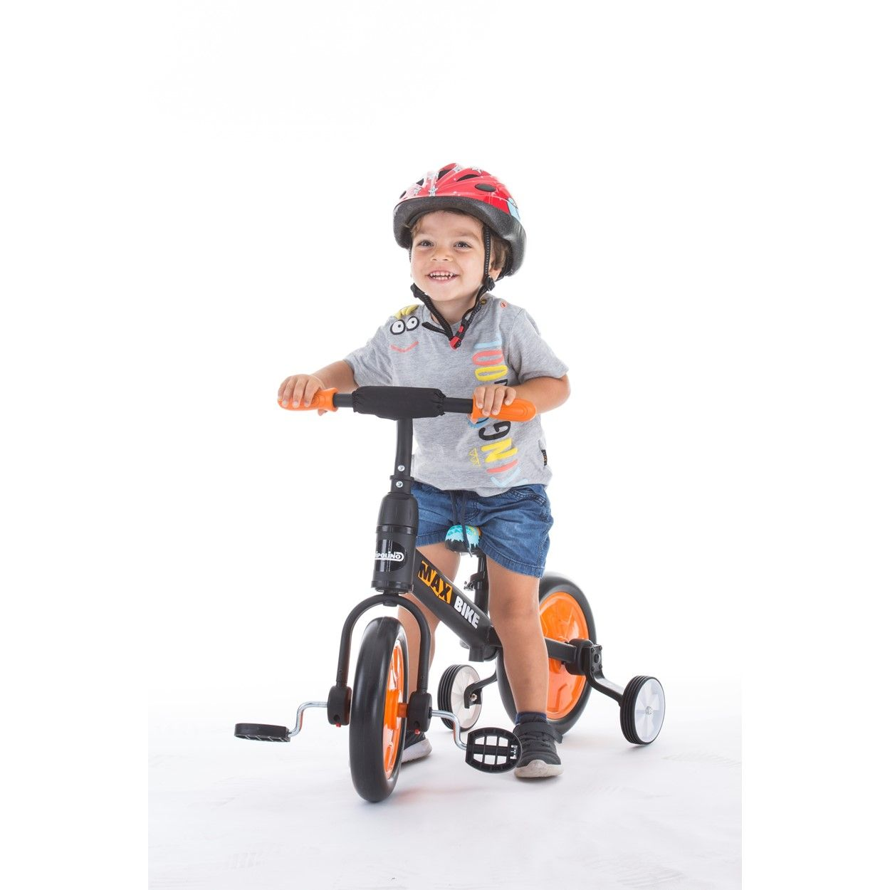 Un niño pequeño en un triciclo negro y naranja con casco rojo sonríe a la cámara.