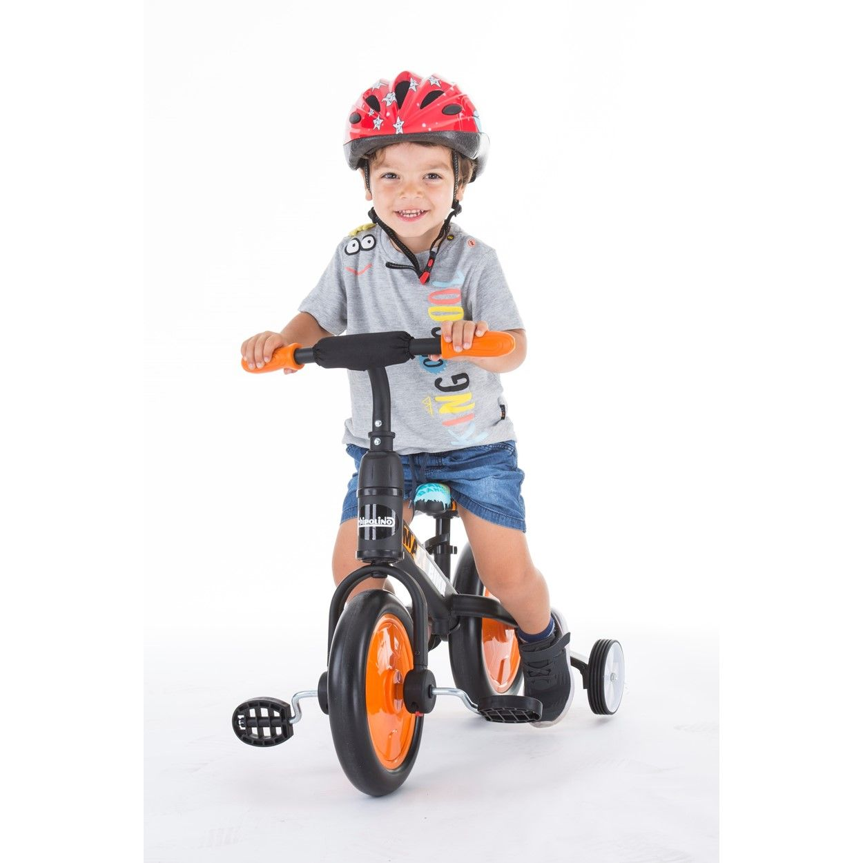 Un niño pequeño con casco monta un triciclo con ruedas y pedales naranjas.
