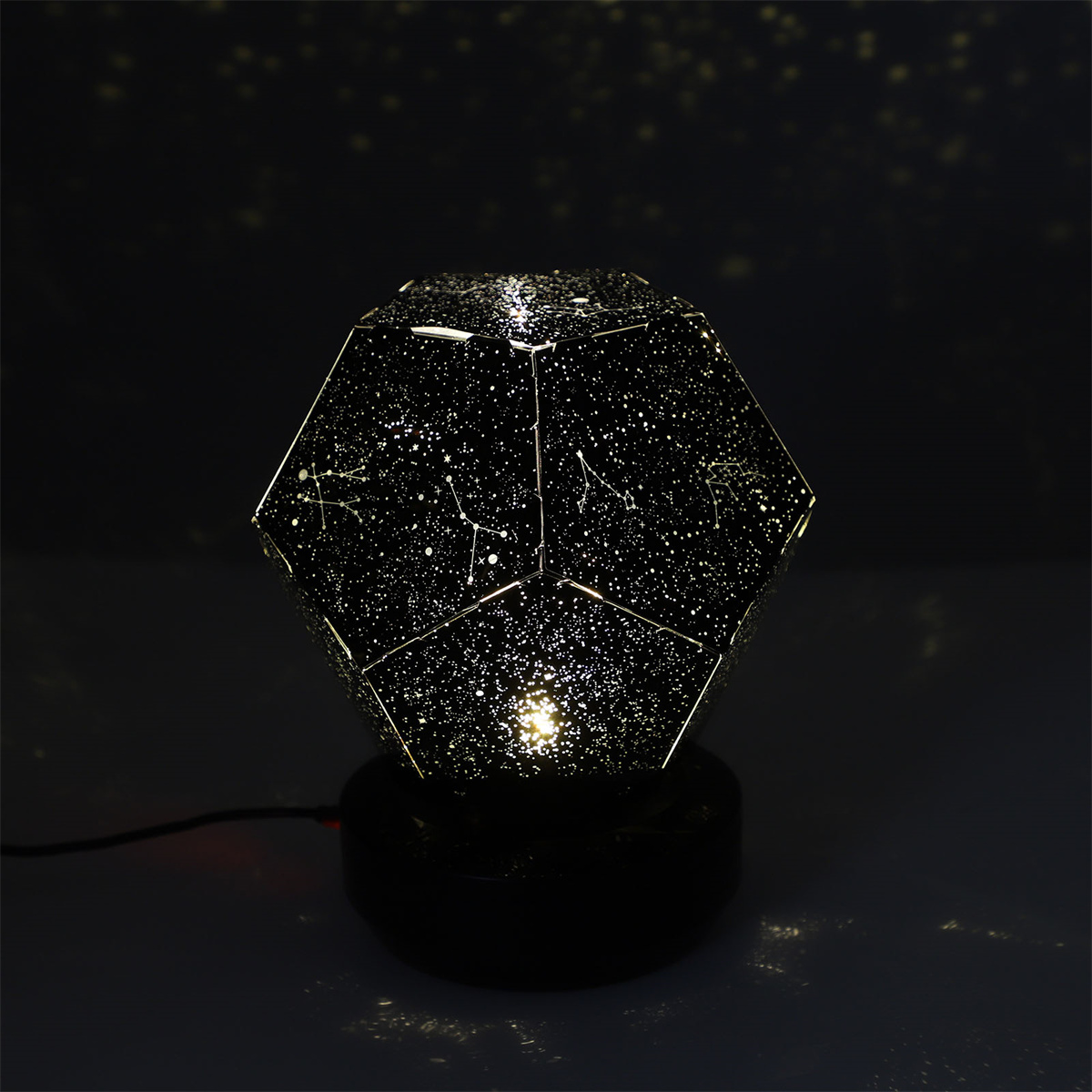 Eine Sternprojektorlampe auf schwarzem Sockel, die Sterne auf eine dunkle Oberfläche projiziert.