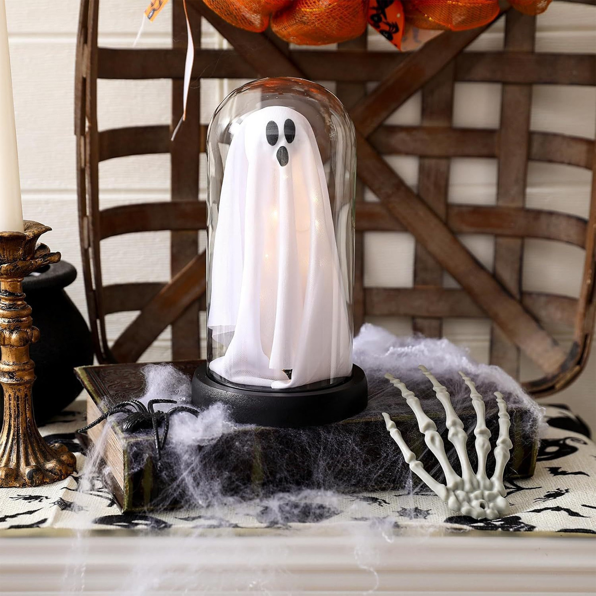 Ein Halloween-Geist aus weißem Stoff in einer Glasglocke. Er sitzt auf einem Buch mit Spinnweben, in der Nähe einer Kerze.