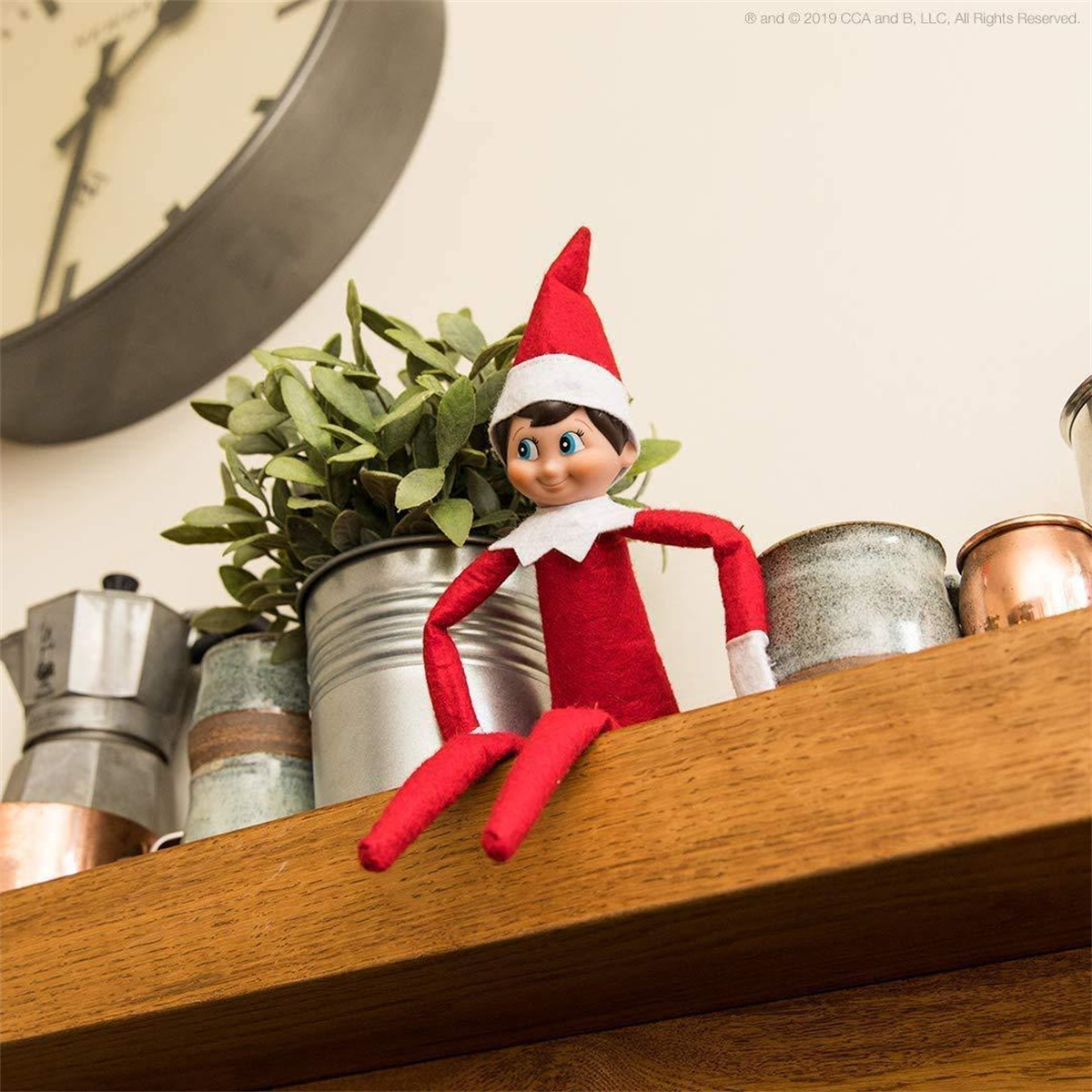 Eine Elf on the Shelf-Puppe ist auf einem Holzregal. Im Hintergrund sind eine Uhr und Pflanzen.