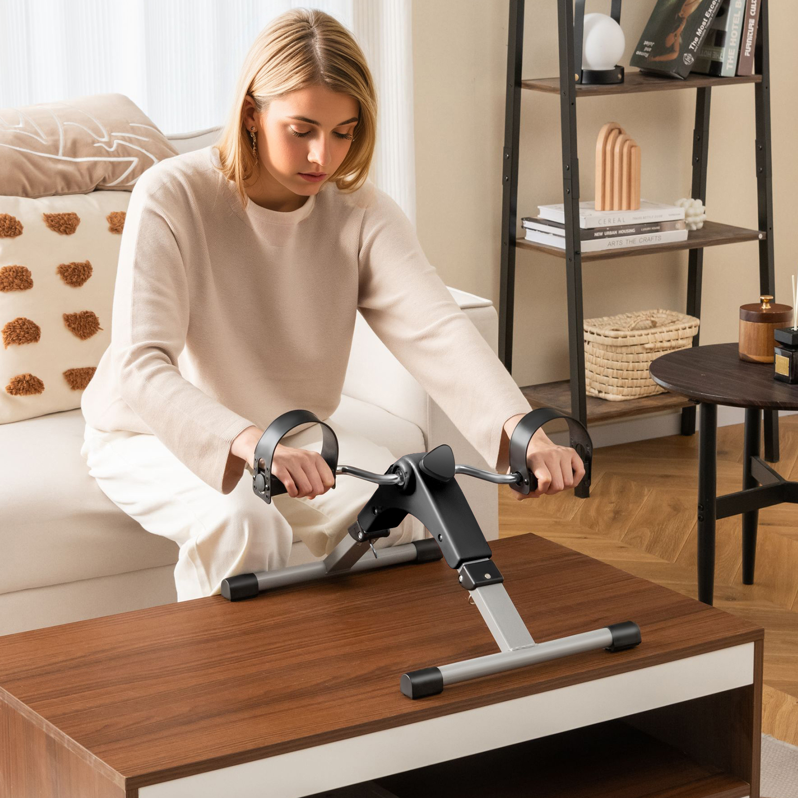 Frau trainiert auf einem Tisch mit einem Pedal-Trainingsgerät, mit Couch und Büchern im Hintergrund.