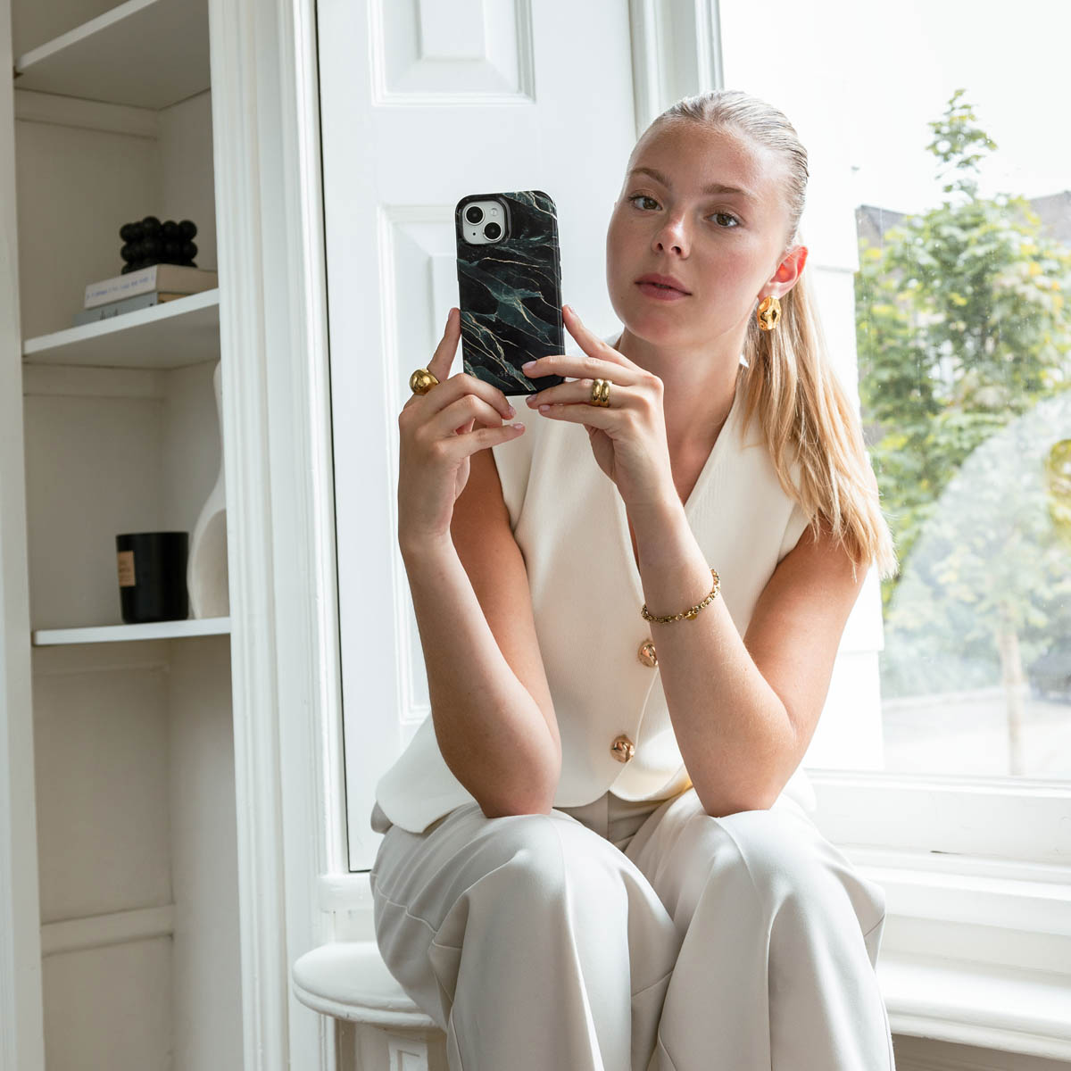 Femme en blanc tenant téléphone avec coque en marbre. Assise près d'une fenêtre blanche.