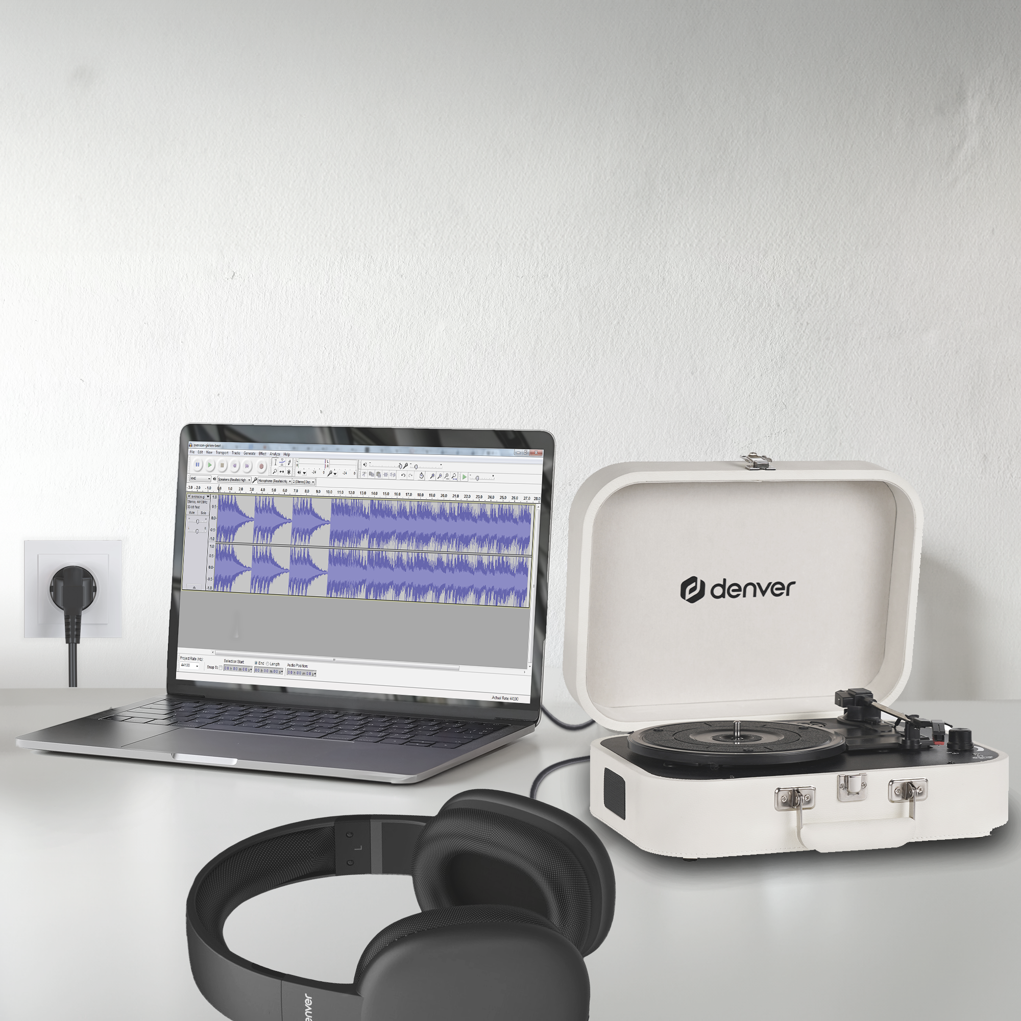 Laptop z otwartym oprogramowaniem do edycji audio obok białego gramofonu i czarnych słuchawek na białym stole.