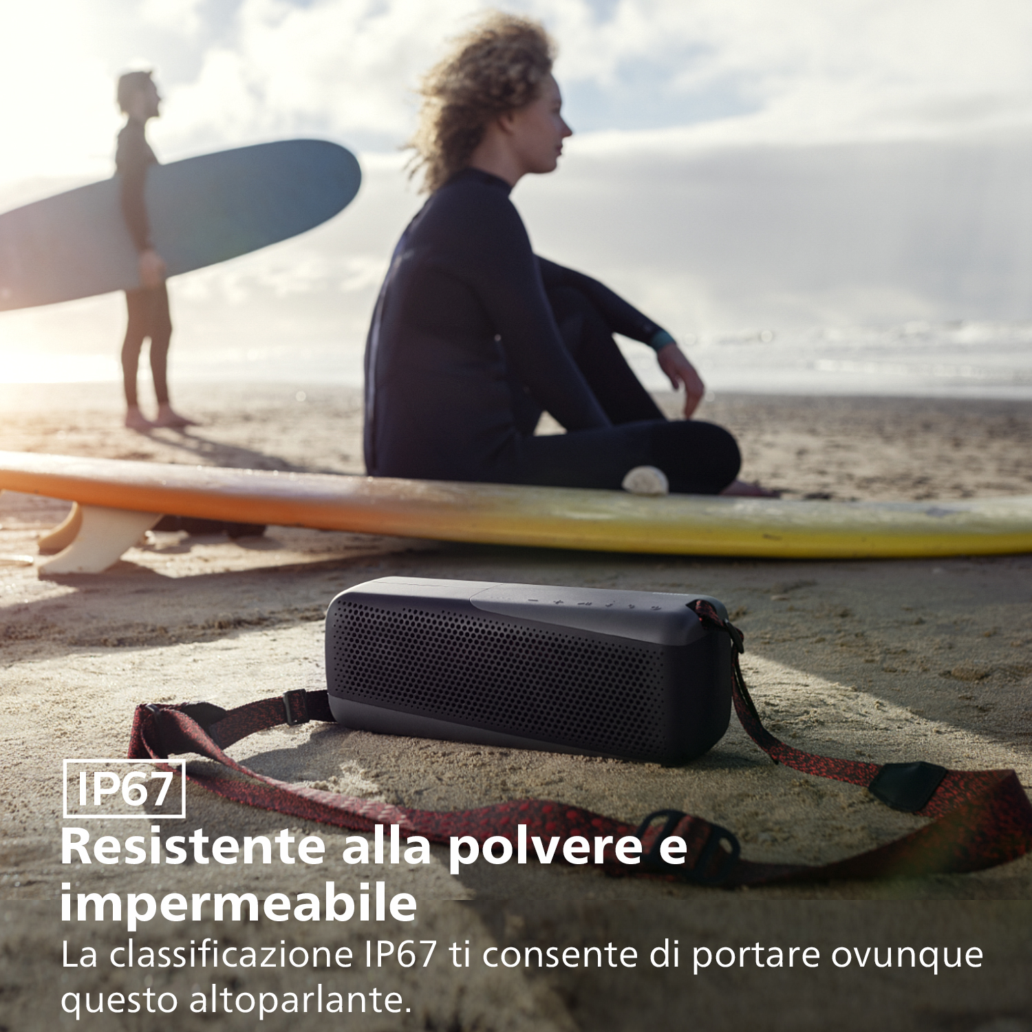 Czarny głośnik przenośny na piaszczystej plaży, z surferami i wodą w tle.