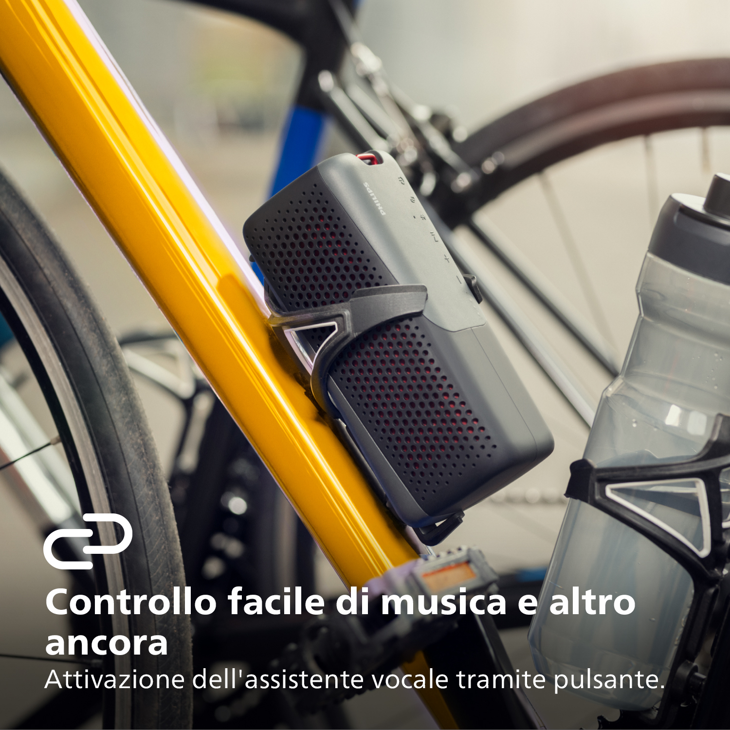 Szary głośnik zamontowany na żółtej ramie roweru. Butelka z wodą w pobliżu. Tekst nałożony.