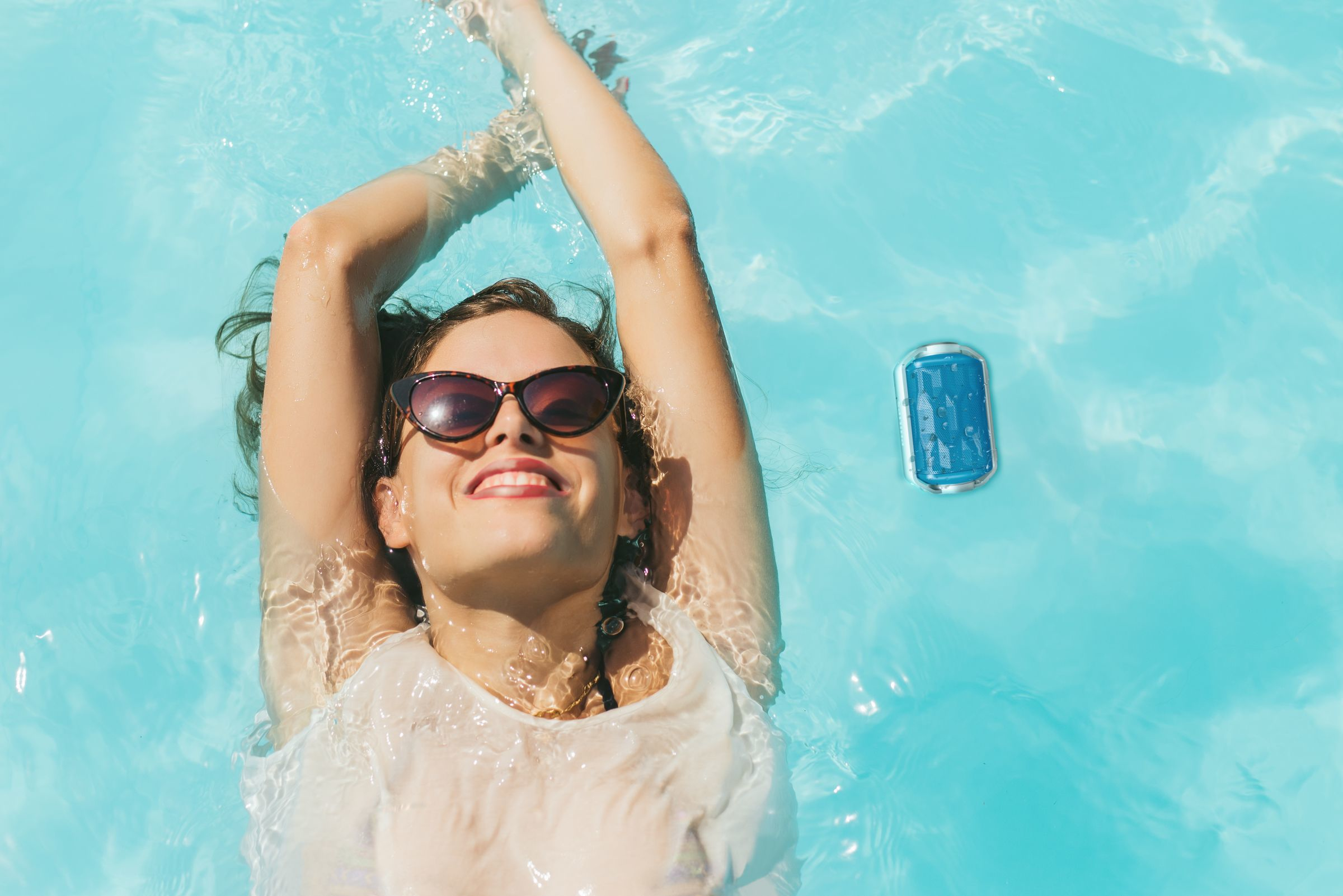 Mujer con gafas de sol en una piscina. Nada, con los brazos levantados. Un altavoz azul flota cerca.