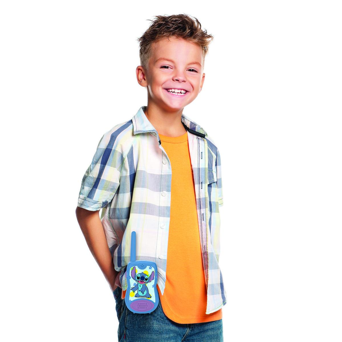Młody chłopiec w koszuli w kratę i pomarańczowej koszulce trzymający krótkofalówkę Stitch. Uśmiecha się.