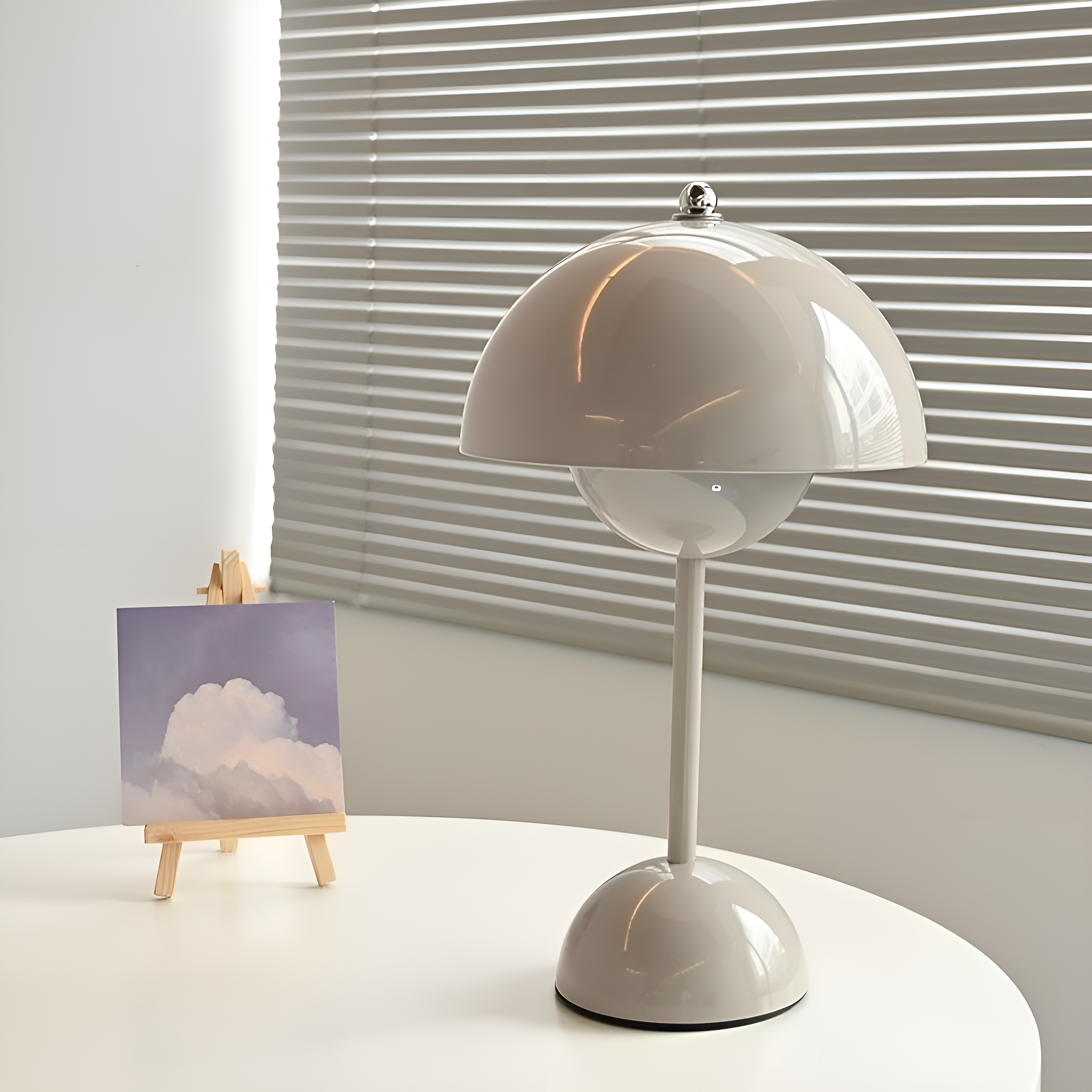 Eine graue Lampe auf einem weißen Tisch, mit einem Gemälde auf einer kleinen Staffelei. Jalousien im Hintergrund.