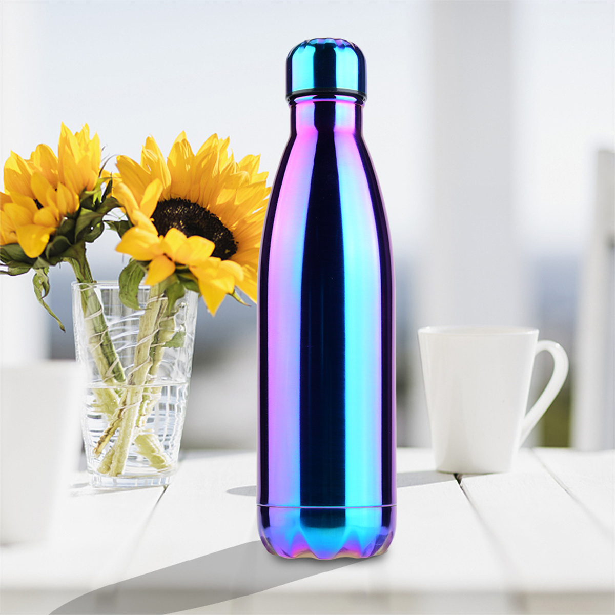 Eine bunte Wasserflasche neben Sonnenblumen in einer Vase und einer weißen Tasse.