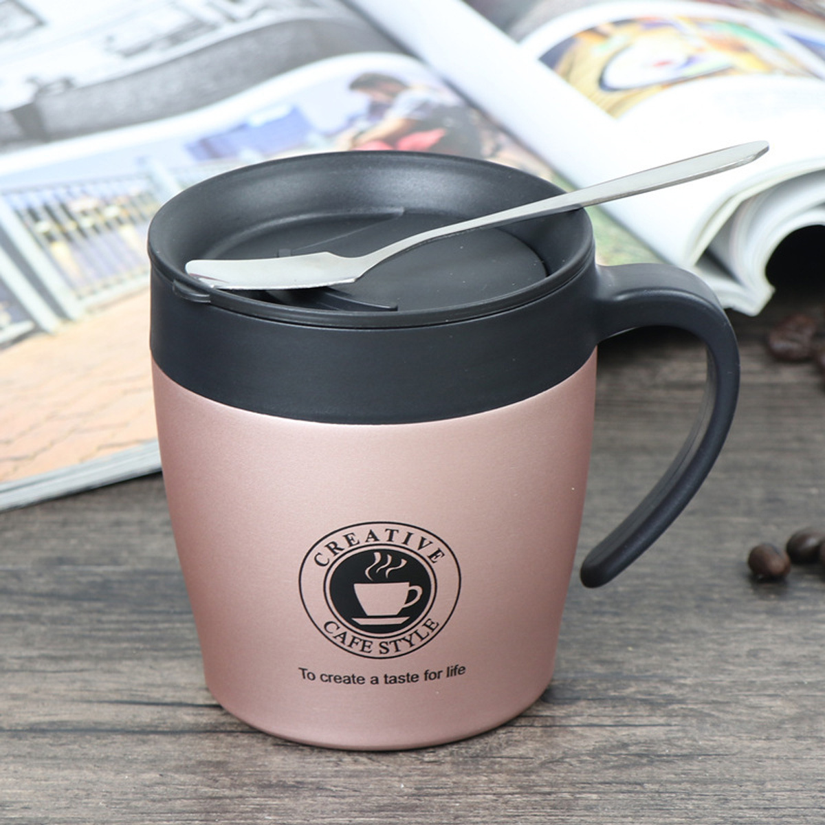 Ein roségoldener Reisebecher mit schwarzem Deckel und Löffel. Auf dem Becher ist das Creative Cafe Style Logo.