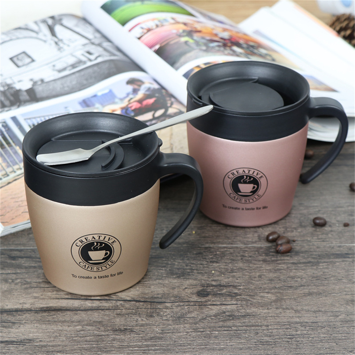 Zwei goldfarbene Tassen, schwarze Griffe und Deckel. Eine hat einen Löffel und "Creative Cafe Style" aufgedruckt.