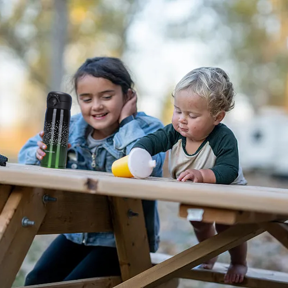 Zwei Kinder an einem Picknicktisch. Ein Mädchen lächelt. Das andere Kind hält eine Flasche.