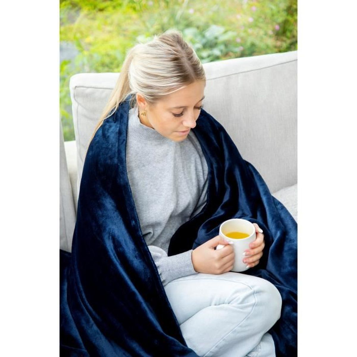 Frau in blauer Decke, hält eine Tasse mit Getränk, sitzt draußen auf einem hellen Sofa.