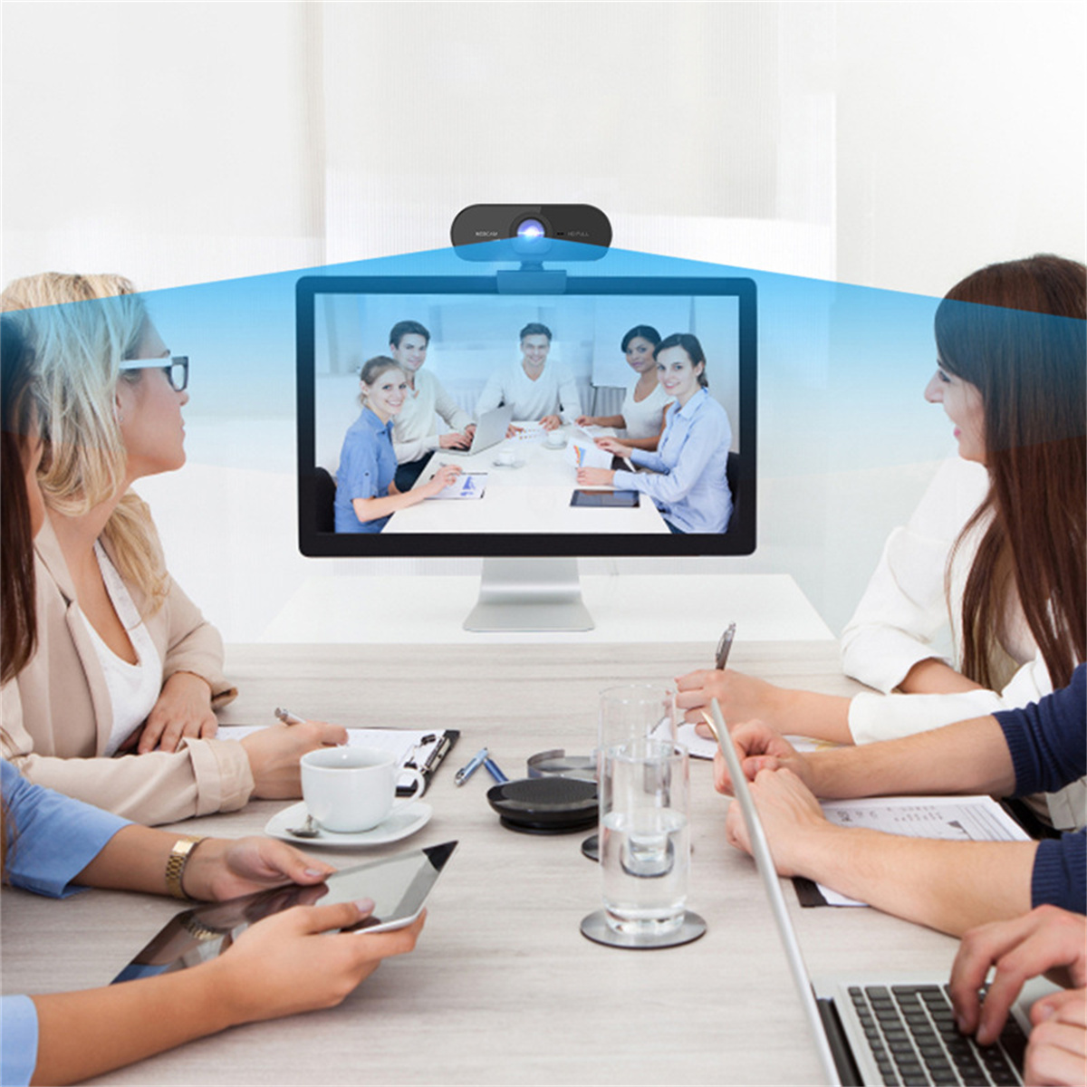 Sitzungssaal mit Webcam. Personen auf dem Monitor sind sichtbar. Teilnehmer um den Tisch.