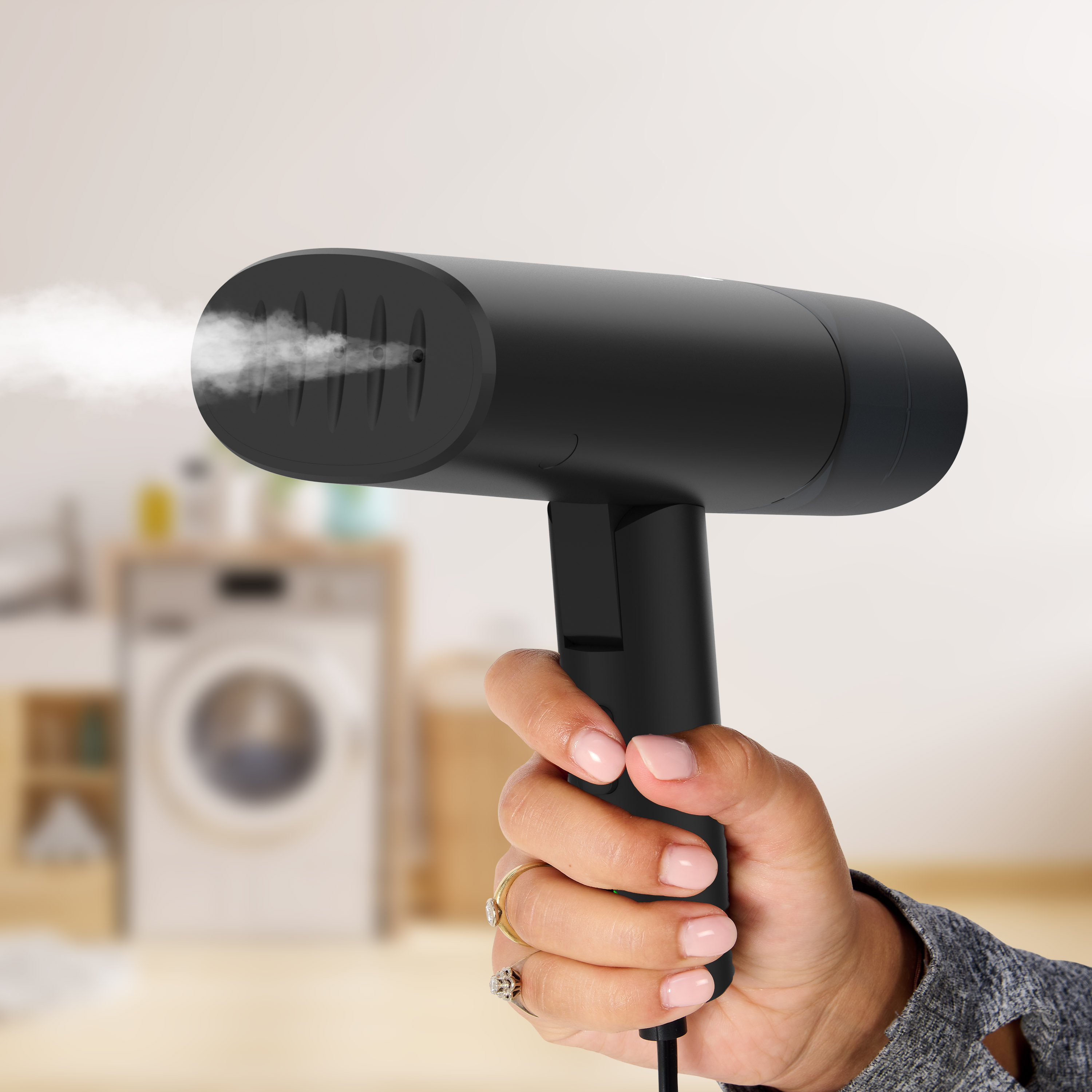 A hand holding a black steamer, releasing steam. Blurred background with laundry.