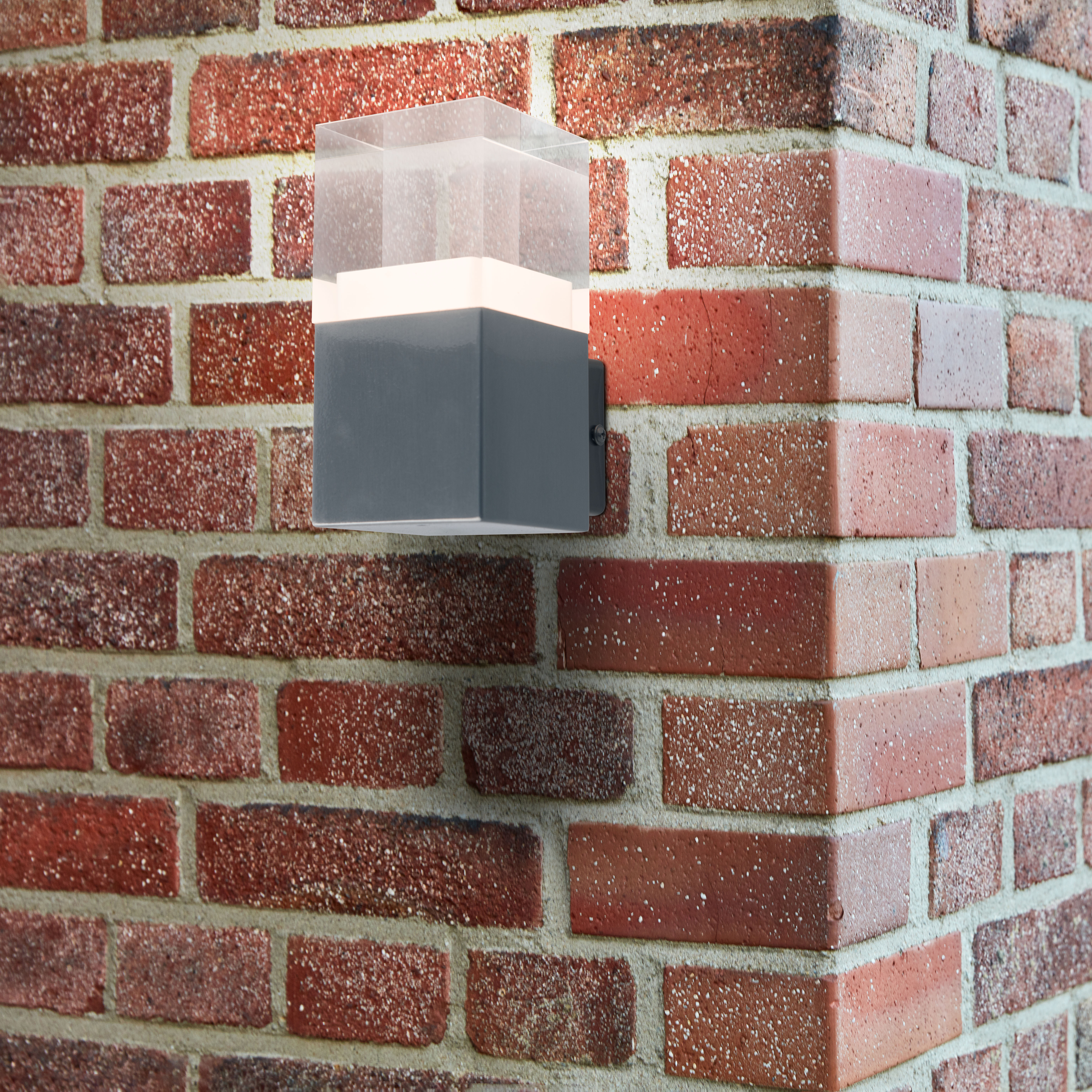 Quadratische Außenwandleuchte an einer roten Backsteinmauer, die Licht aussendet.