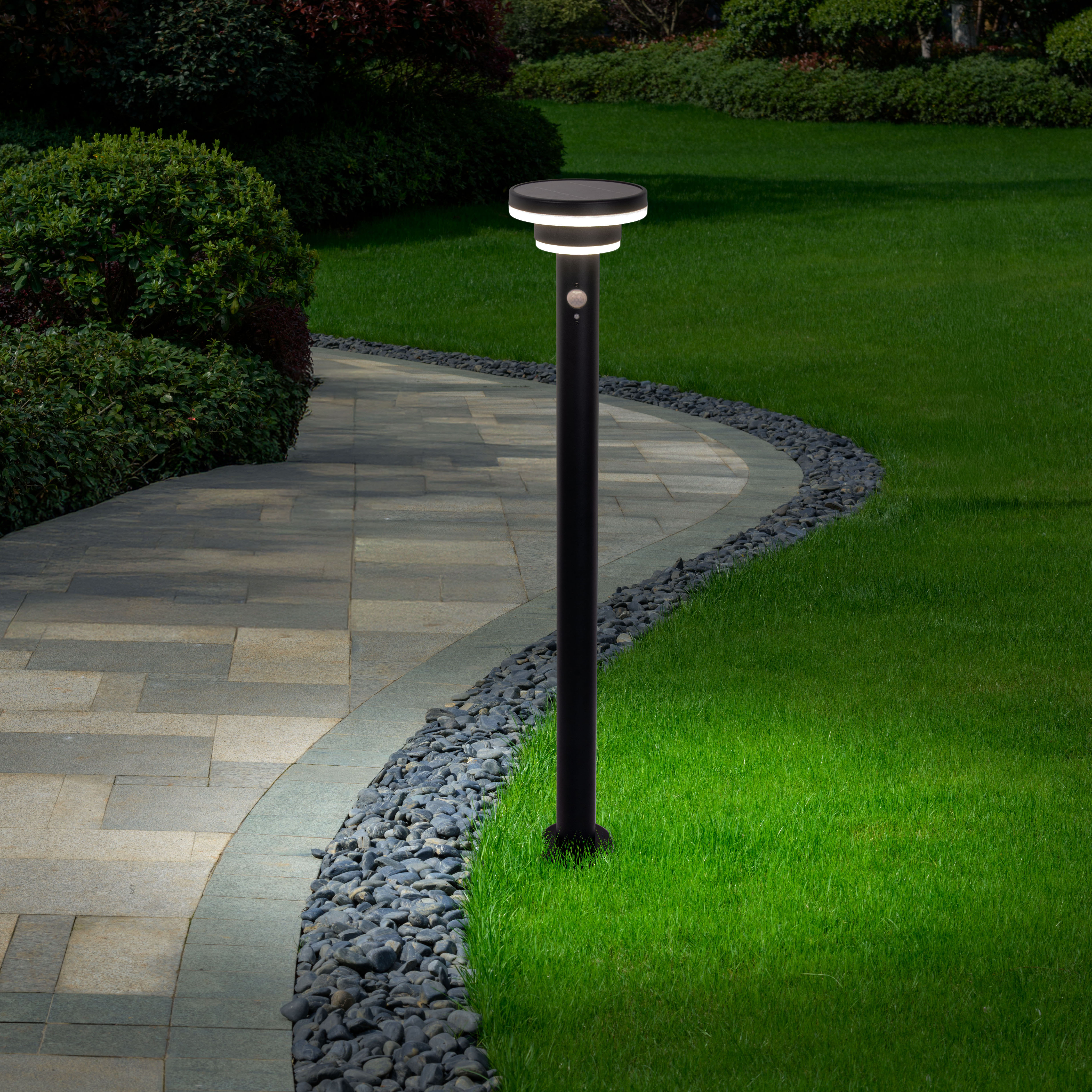 Eine schwarze Außenlampe mit kreisförmigem Licht, die nachts in einem Garten installiert ist. Ein gepflasterter Weg ist in der Nähe.