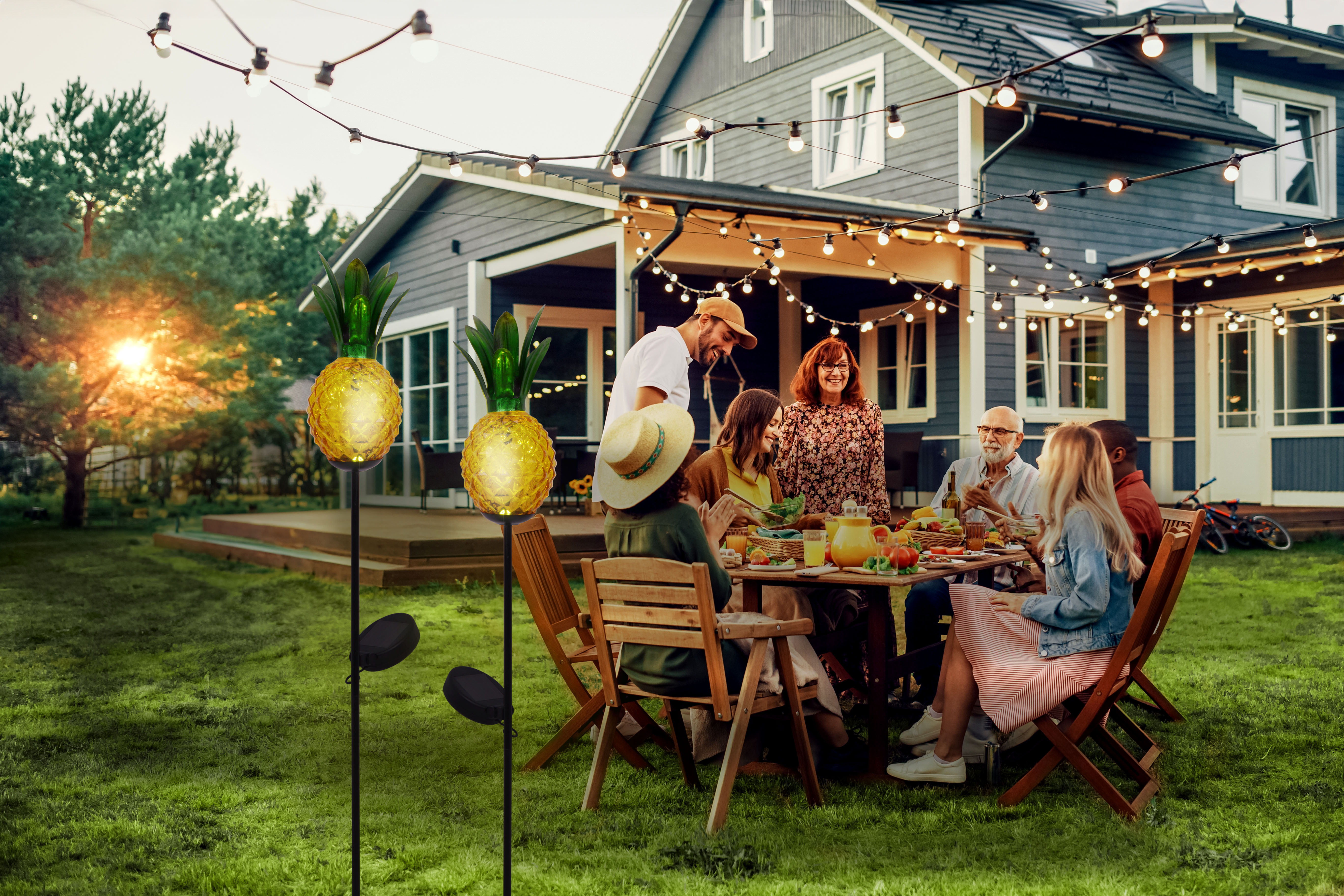 Menschen speisen im Freien an einem Tisch, mit Ananas-Lichtern und einem Haus im Hintergrund.