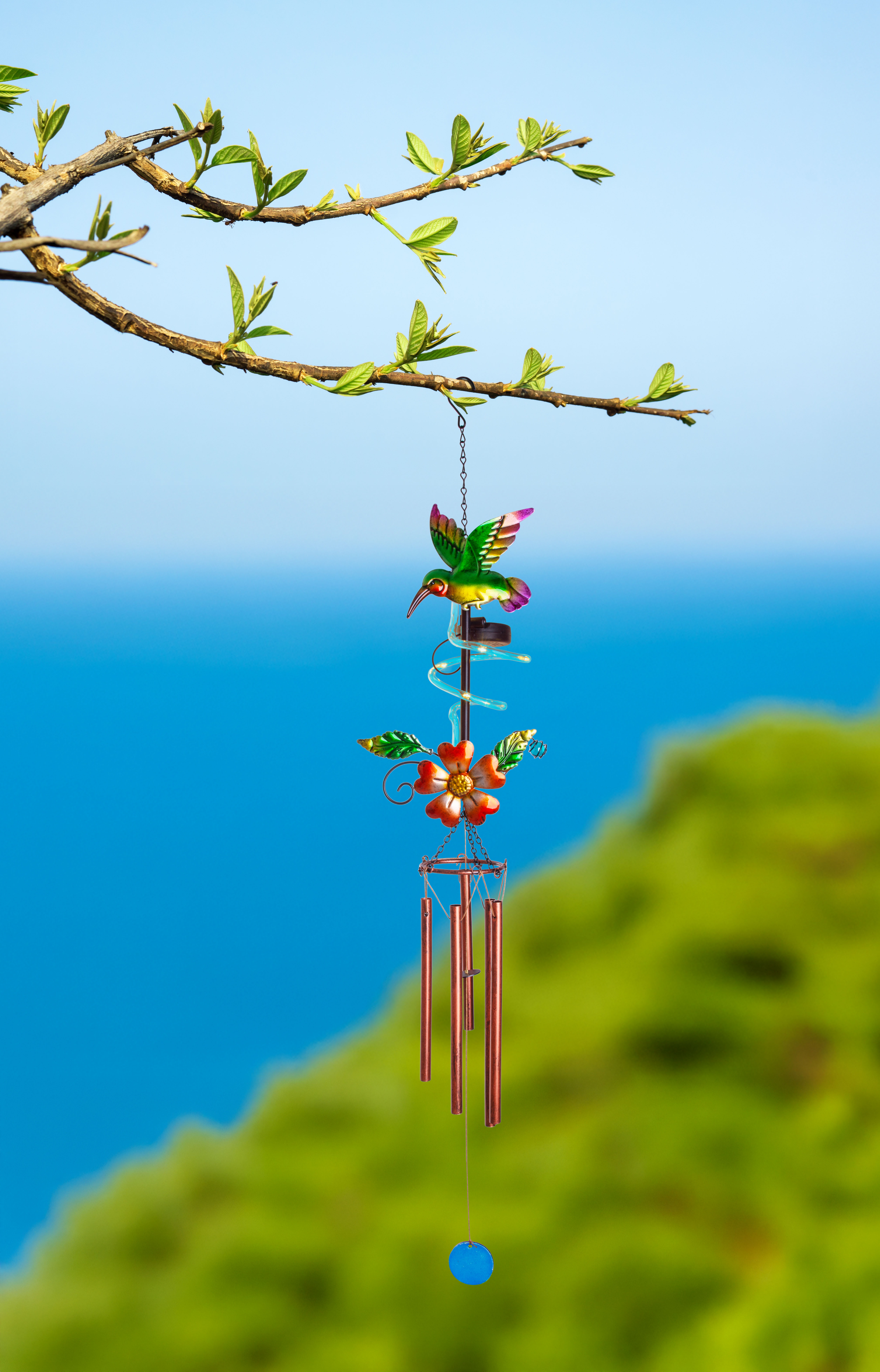 Kolibri-Windspiel, hängt am Ast, blauer Himmel und grünes Laub.