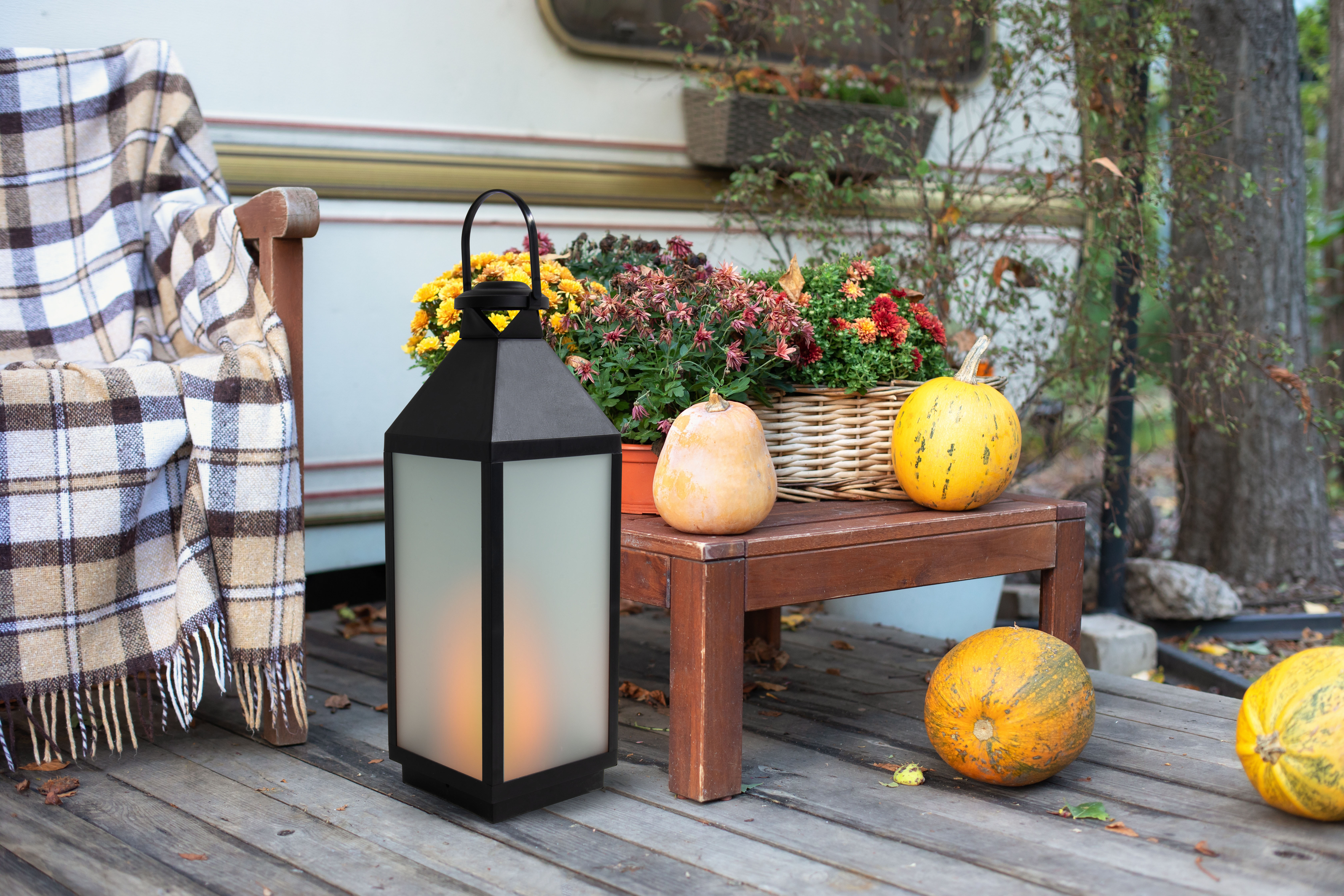 Herbstszene mit Laterne, Kürbissen, Blumen und Decke auf Holzveranda, neben Wohnwagen.