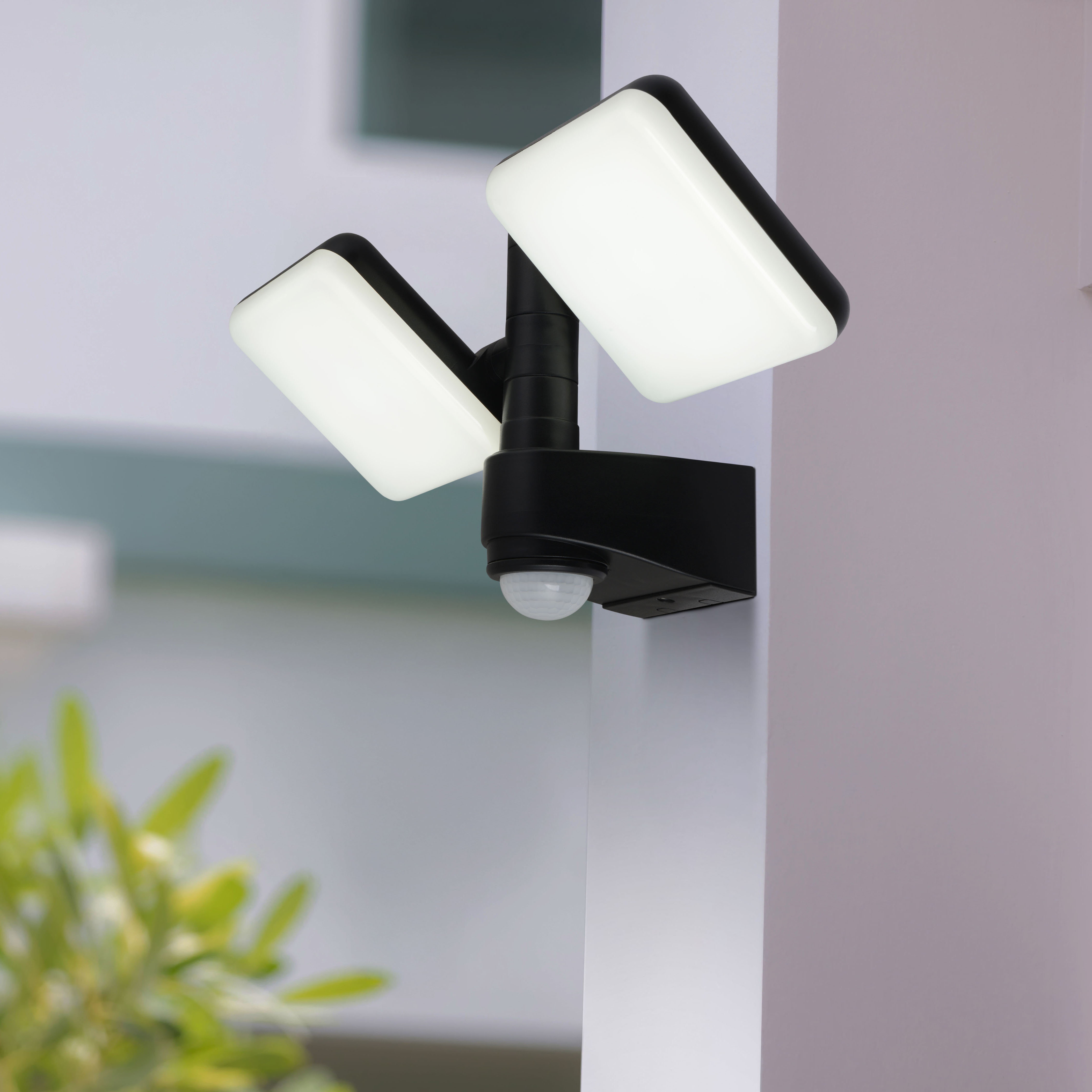 Schwarzer Flutlichtstrahler mit zwei weißen Lichtern, an einer Wand montiert. Ein Sensor ist unten.