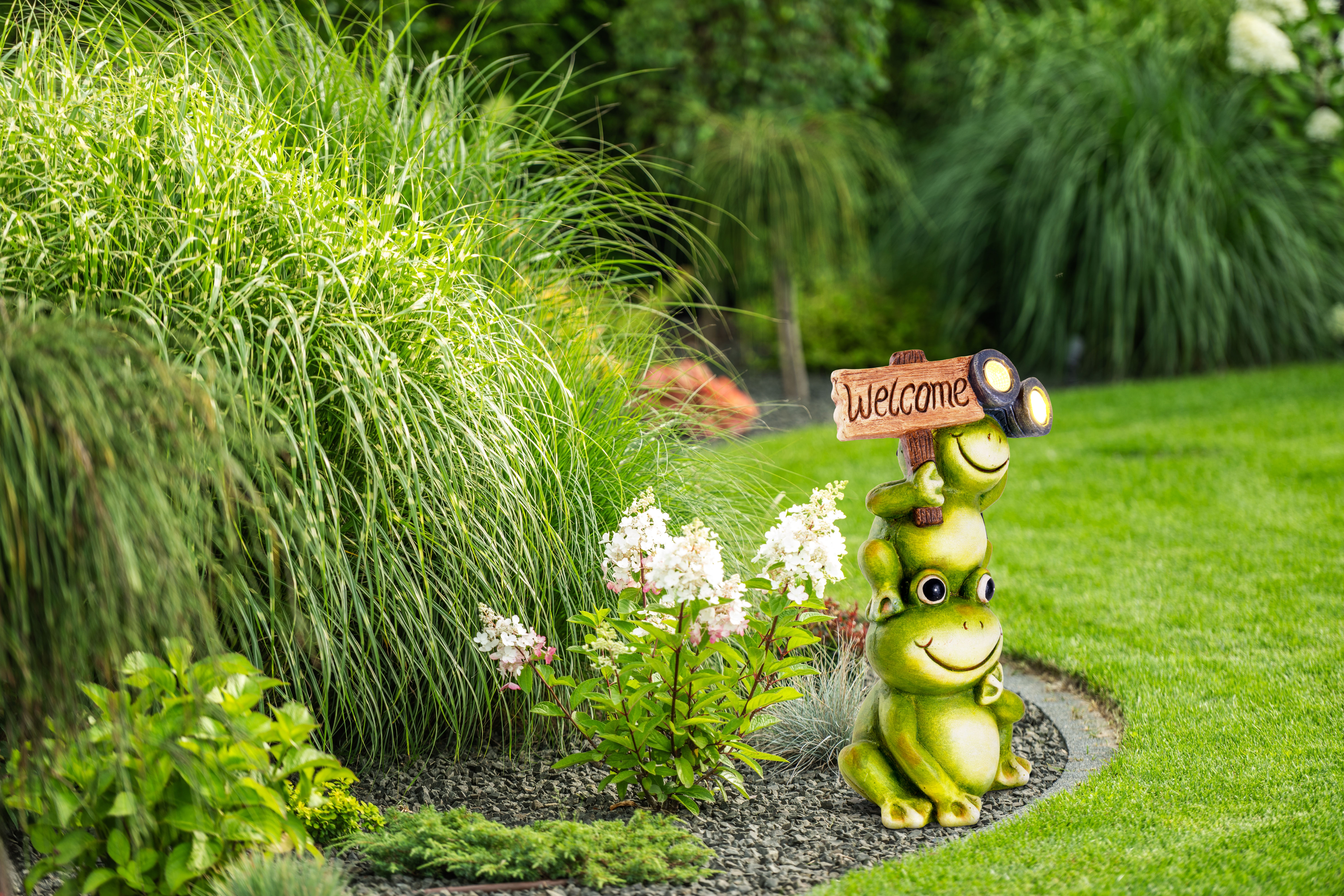 Gartendekoration mit drei Froschfiguren und einem Willkommensschild, in einer Gartenumgebung.