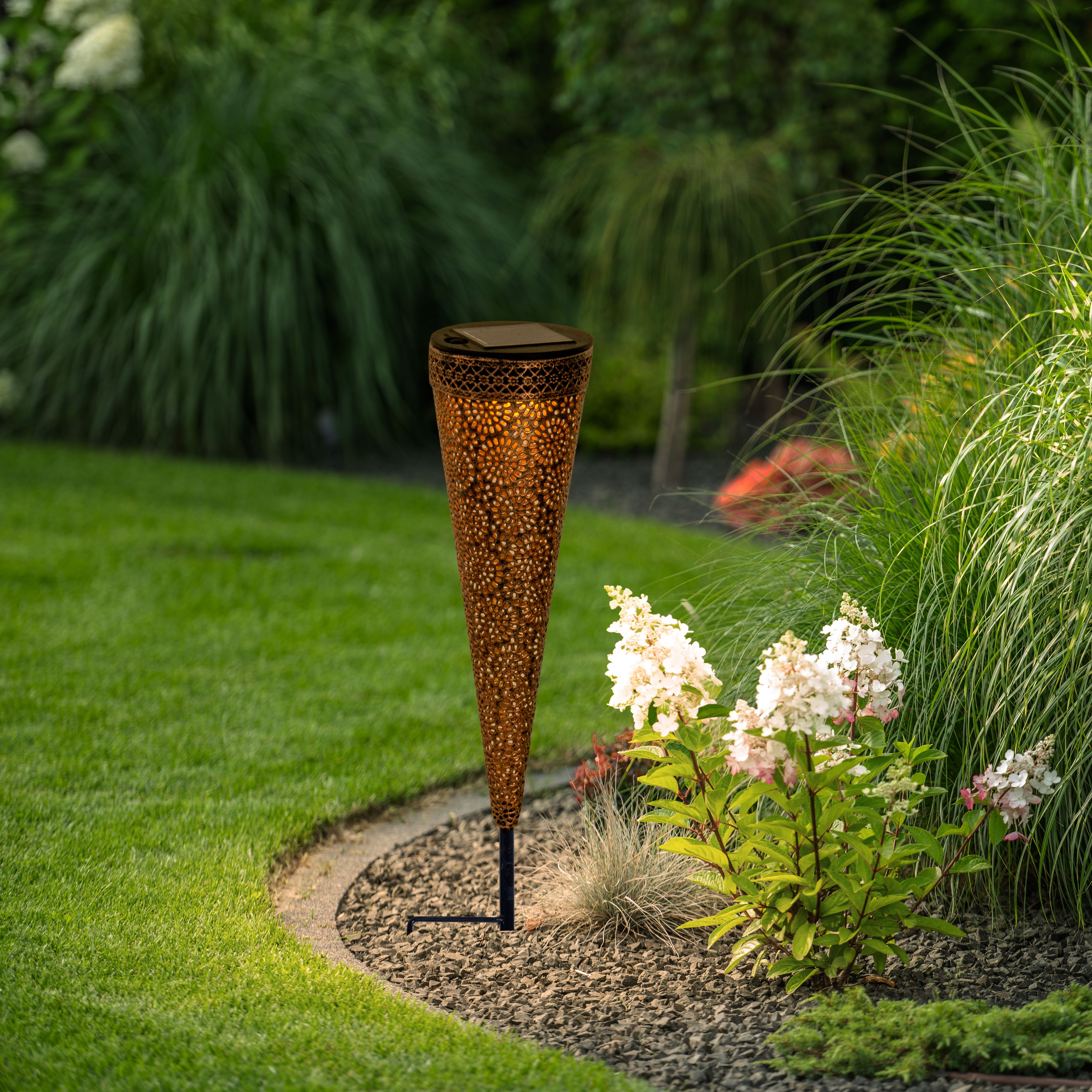 Bronzefarbene Solar-Gartenleuchte in einem Garten mit Gras, Blumen und Grün.