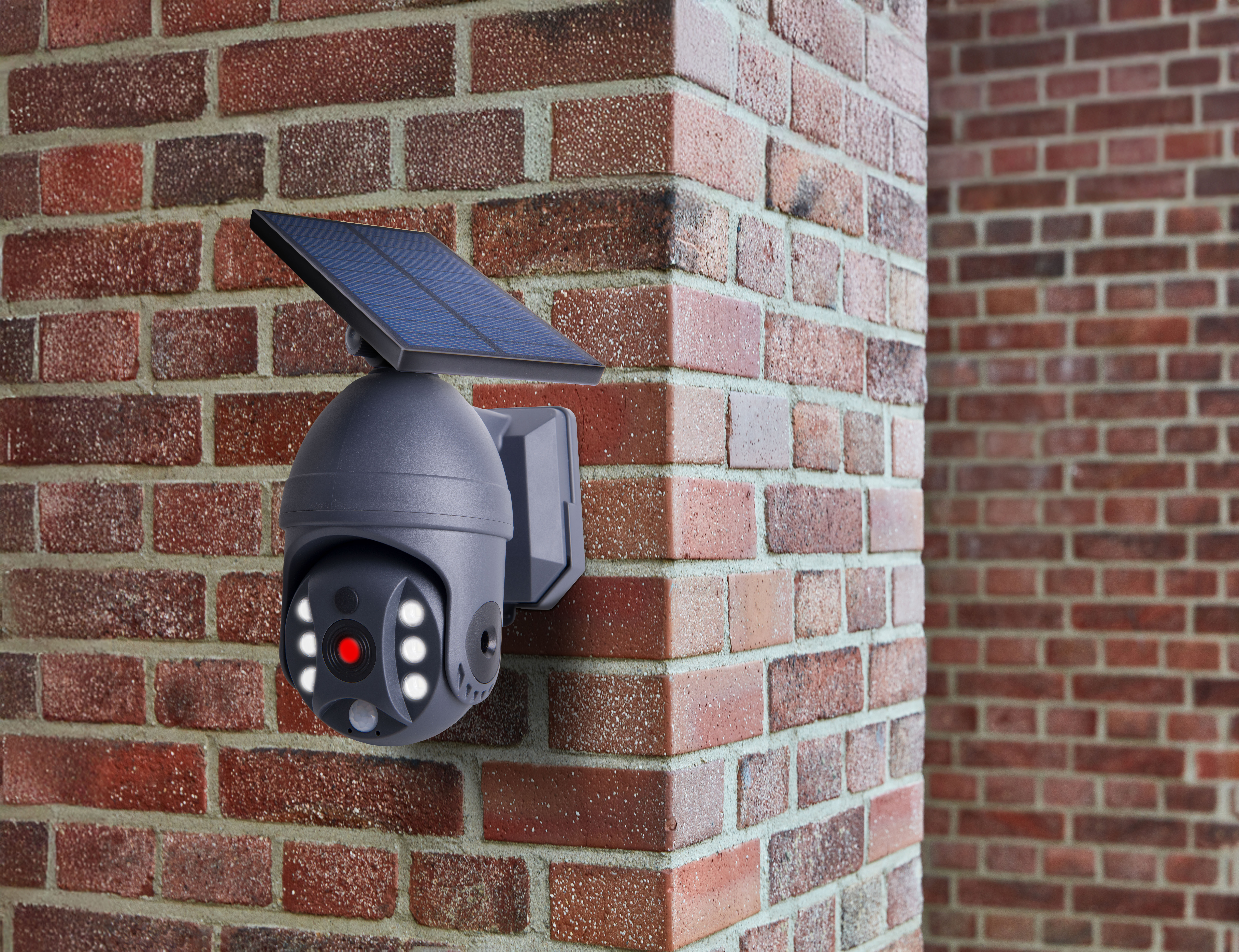 Eine Überwachungskamera, die mit einem Solarpanel an einer Ziegelmauer befestigt ist. Die Kamera hat Lichter und eine rote Linse.