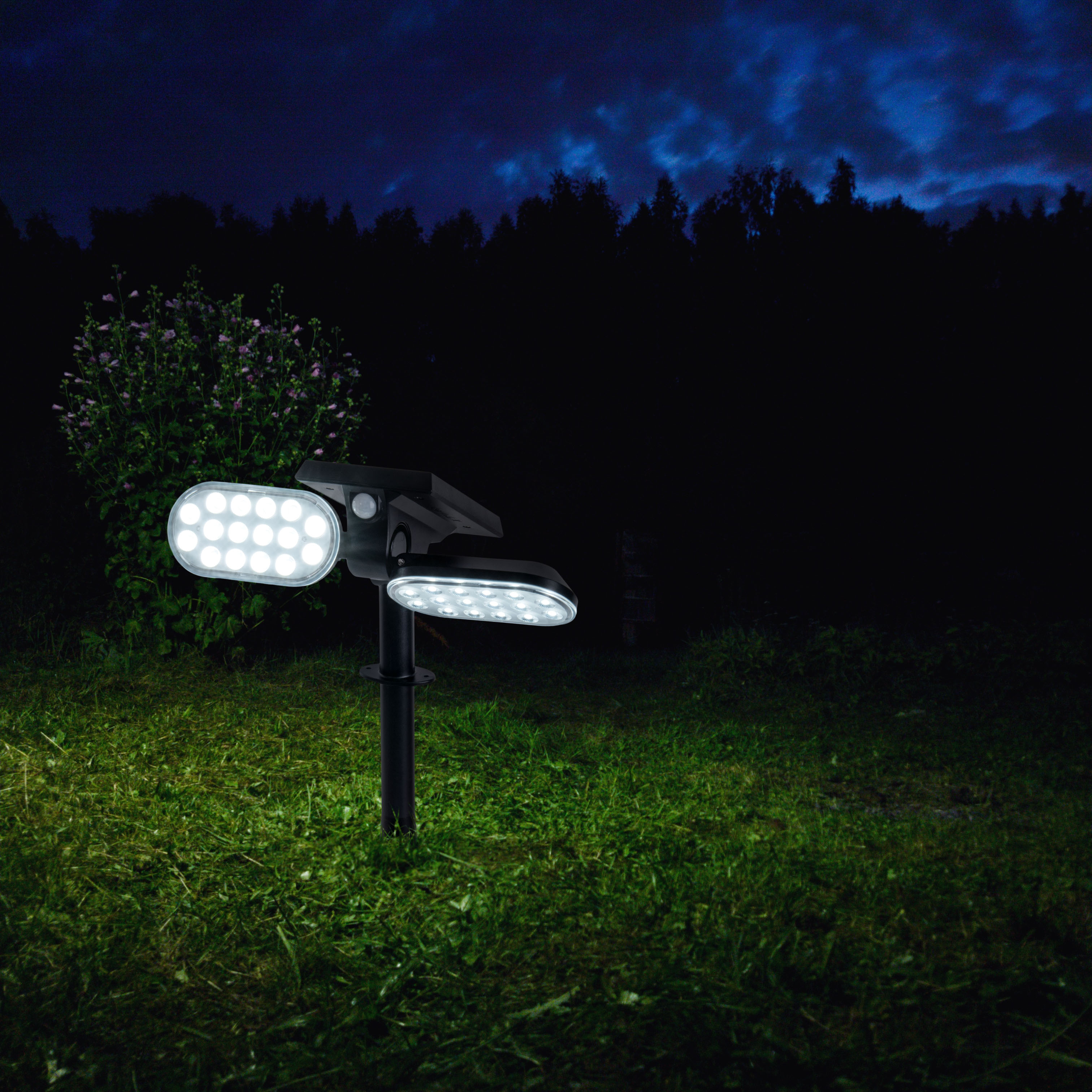 Ein schwarzes Außenlicht mit zwei Lichtpaneelen und einem Solarpanel oben, das nachts in einem Grasbereich aufgestellt ist.