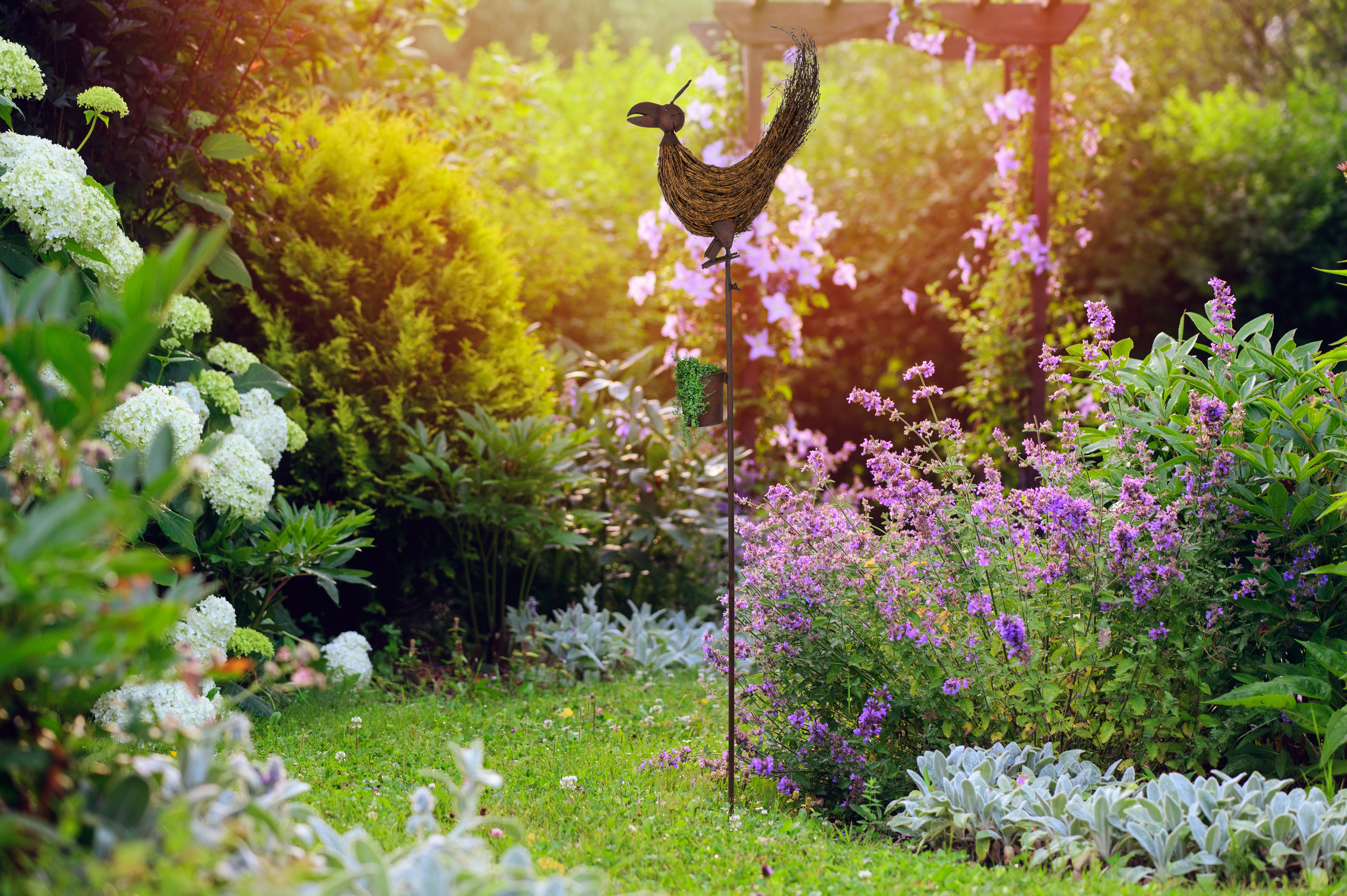 Garten mit einer Metallvogelskulptur, Blumen und Sträuchern. Helles Sonnenlicht.