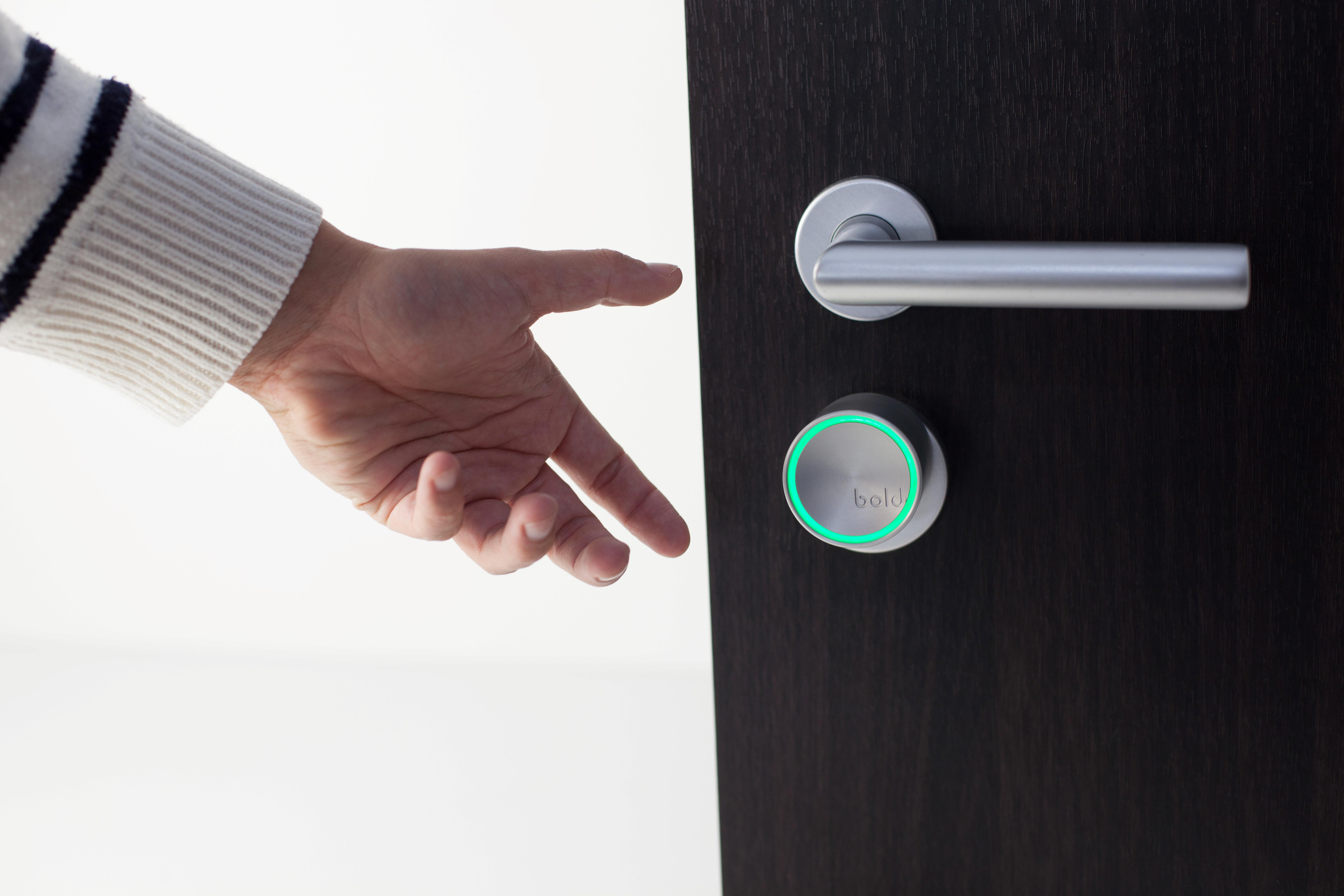 La mano de una persona se acerca a una cerradura inteligente en una puerta de madera oscura. La cerradura brilla en verde.