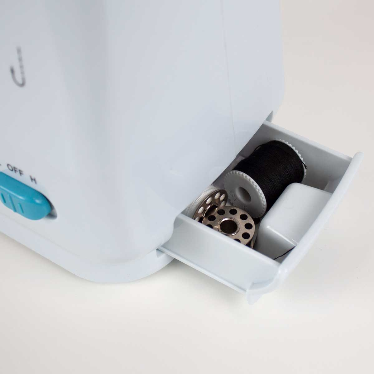 Biała maszyna do szycia z otwartą szufladą. Szuflada zawiera szpulki nici i inne małe przedmioty.